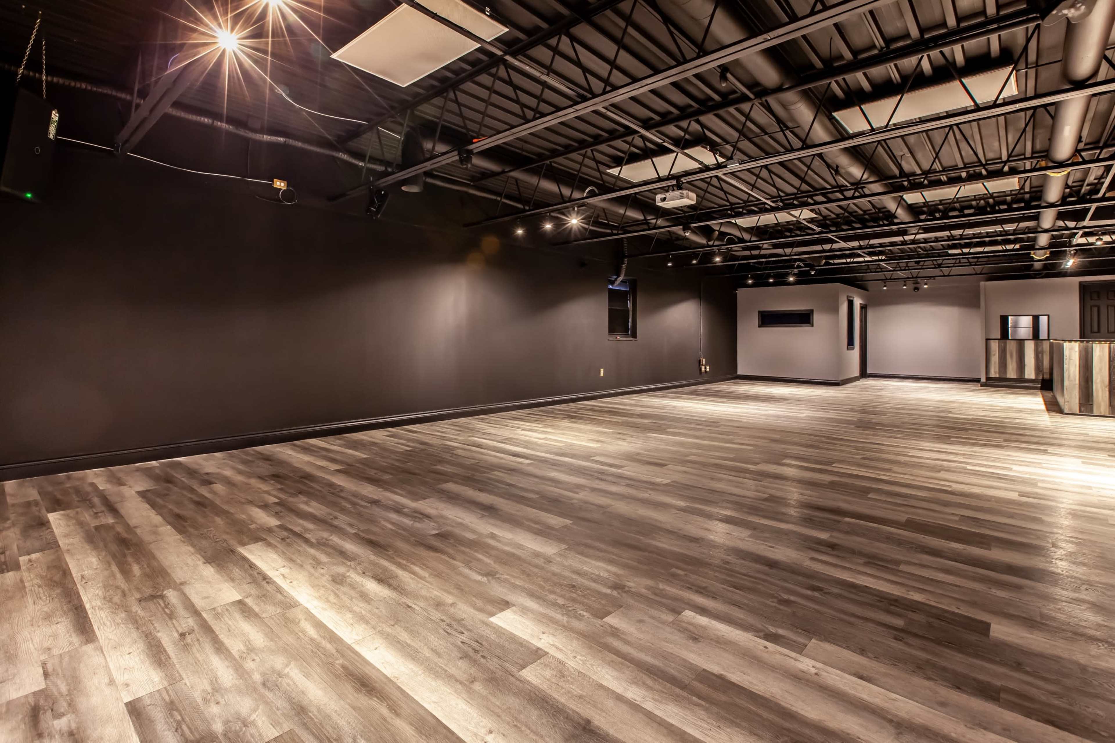 A spacious, empty commercial interior with dark walls and a hardwood floor, illuminated by overhead lighting.