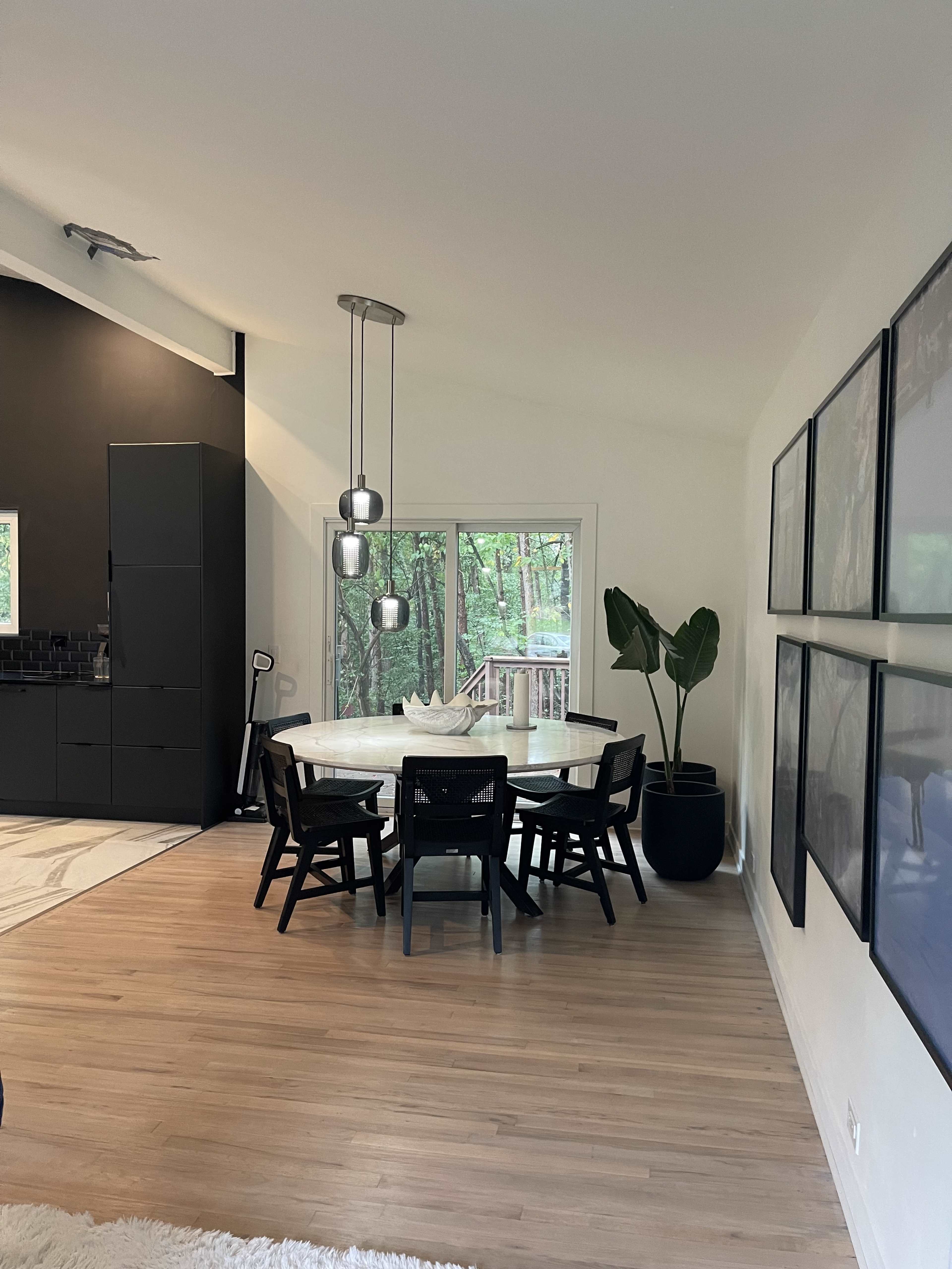 A modern dining area features a round table surrounded by black chairs, with large windows overlooking a wooded view and framed artworks on the wall.