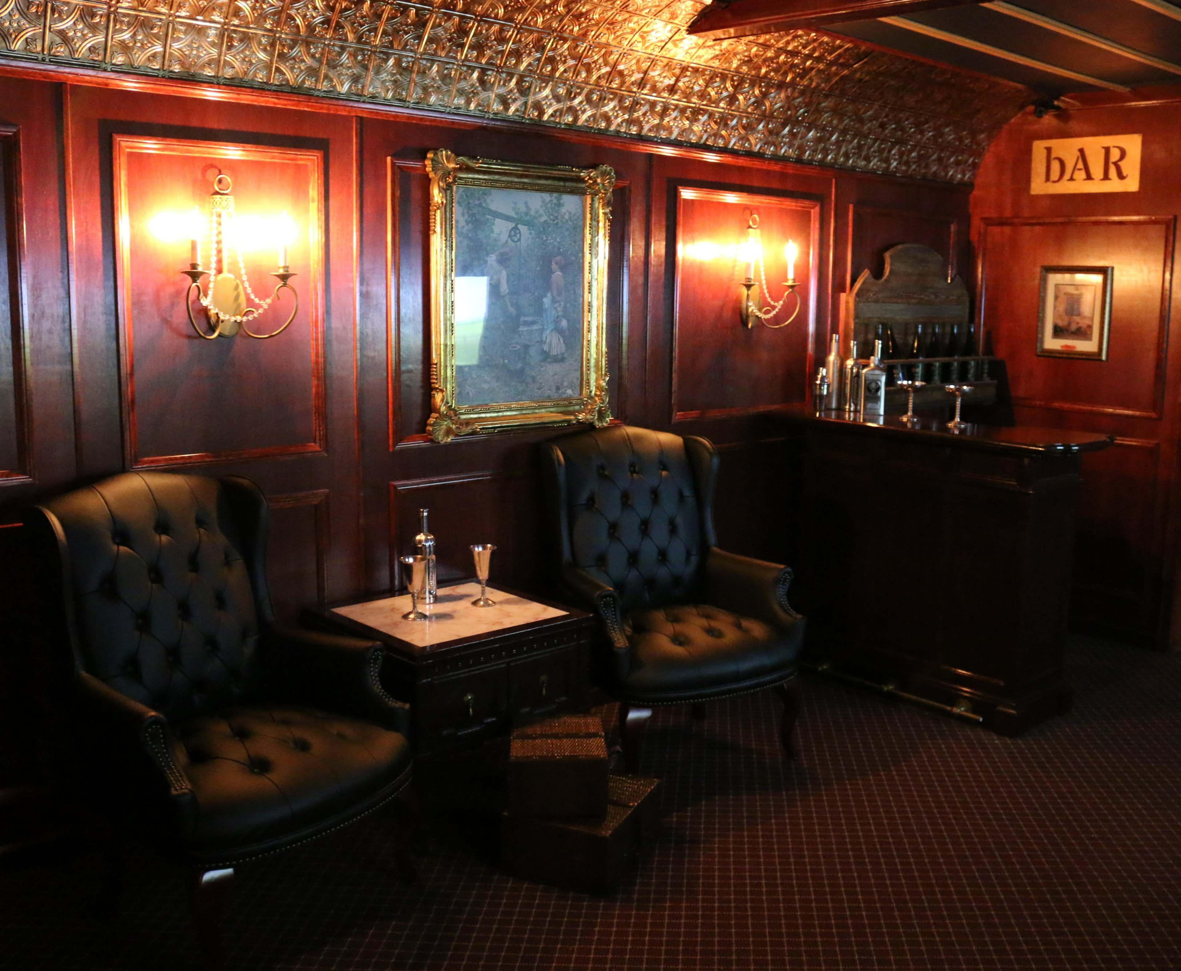 The image shows a wooden-paneled room with two black leather armchairs, a small table between them, and a bar counter in the background.