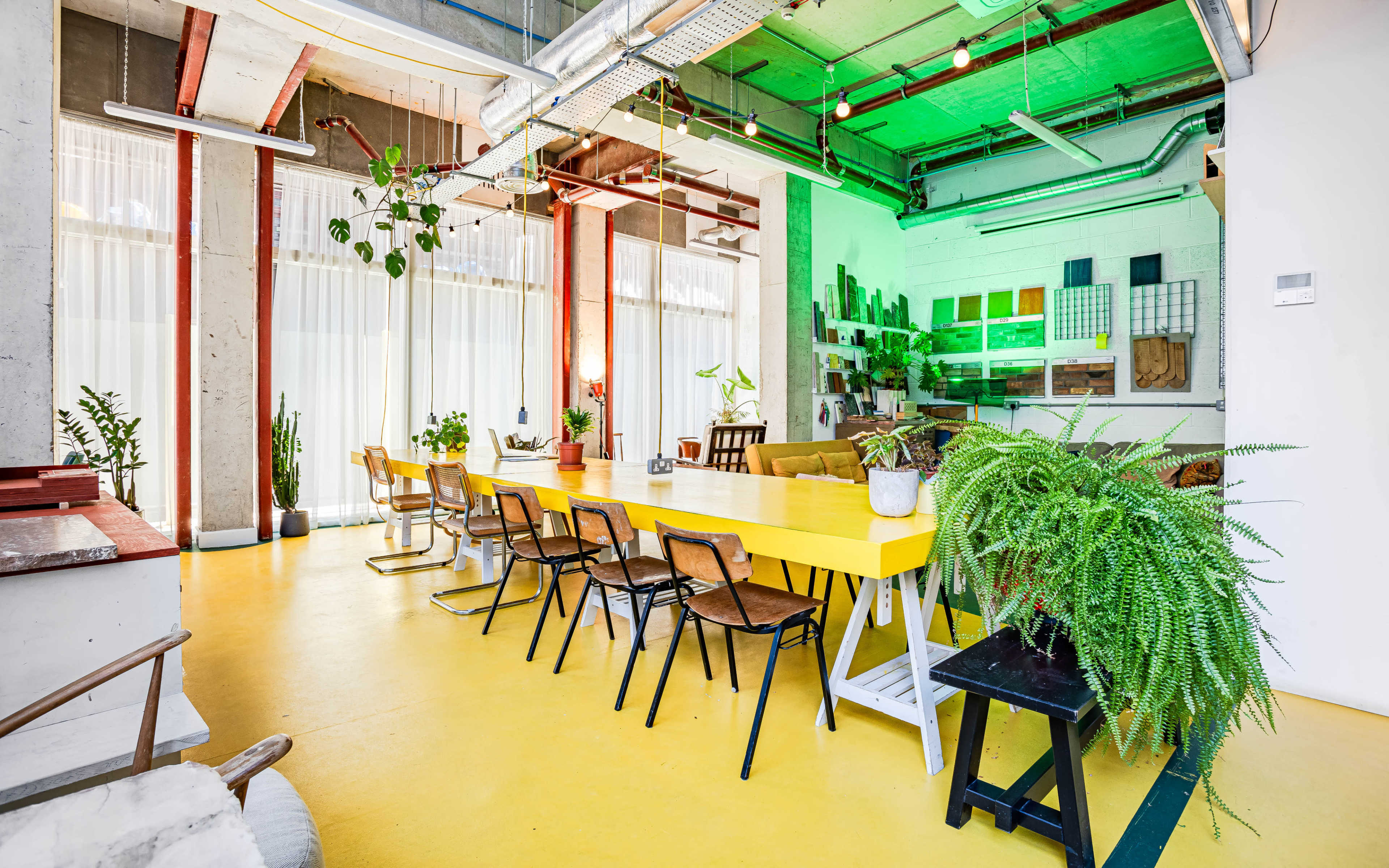 The image shows a bright, modern co-working space featuring a long yellow table surrounded by wooden chairs, plants, and accent lighting.