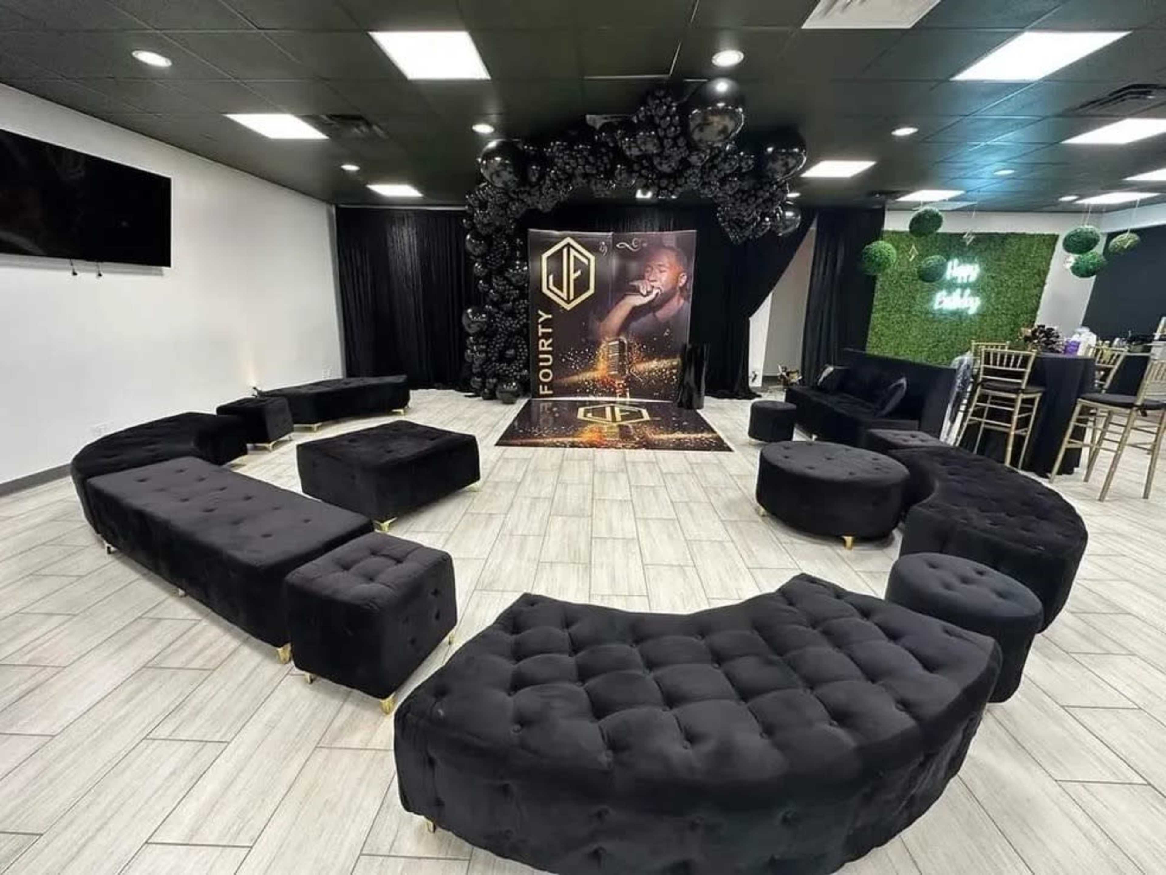 The image shows a stylish event space featuring a large black backdrop with a logo, surrounded by curved black seating and decorative elements.