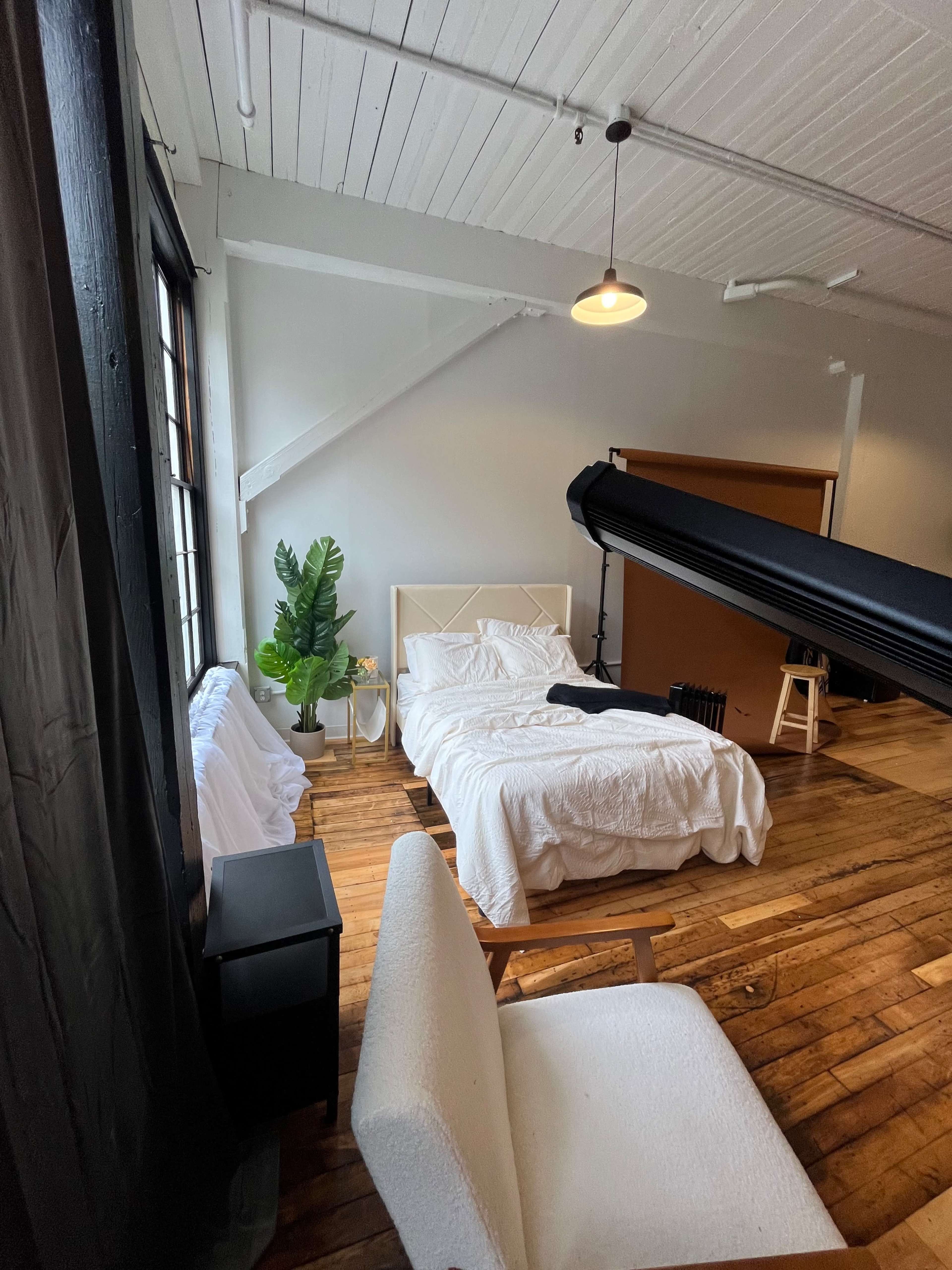 A bedroom with a white bedspread, a bedside table, a chair, and a large window, complemented by a potted plant and wooden flooring.