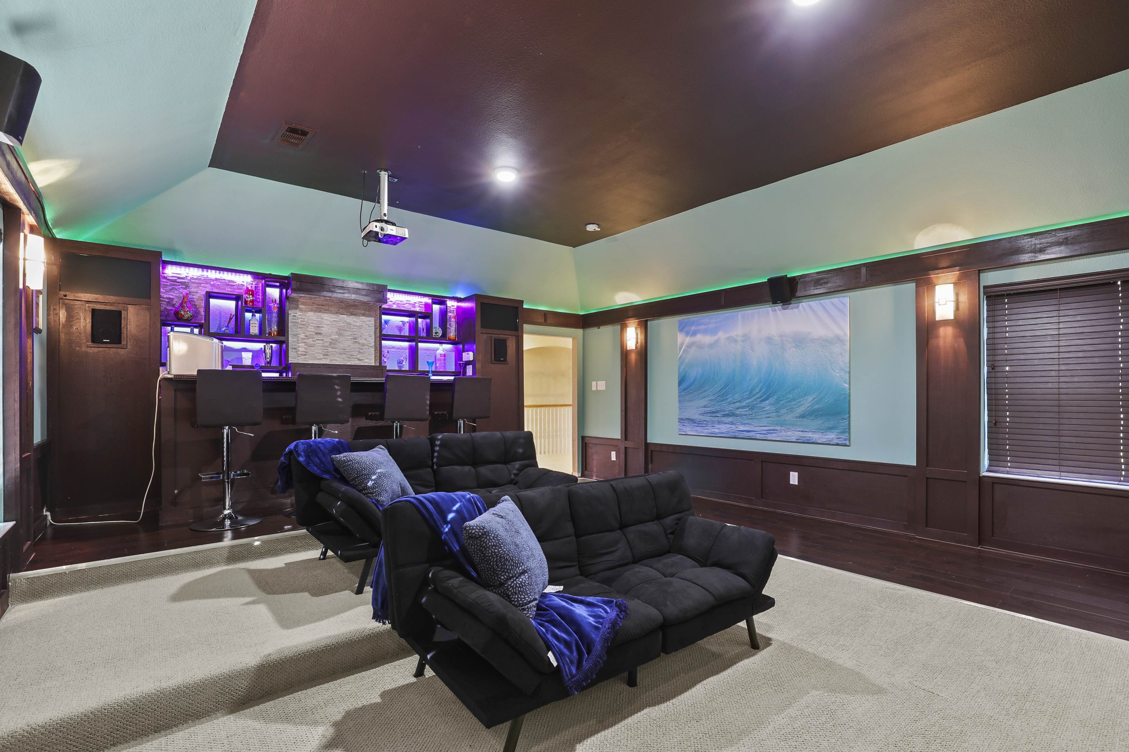 A home theater room featuring black sofas, a bar area with stools, and a large projector screen against a colorful wall.