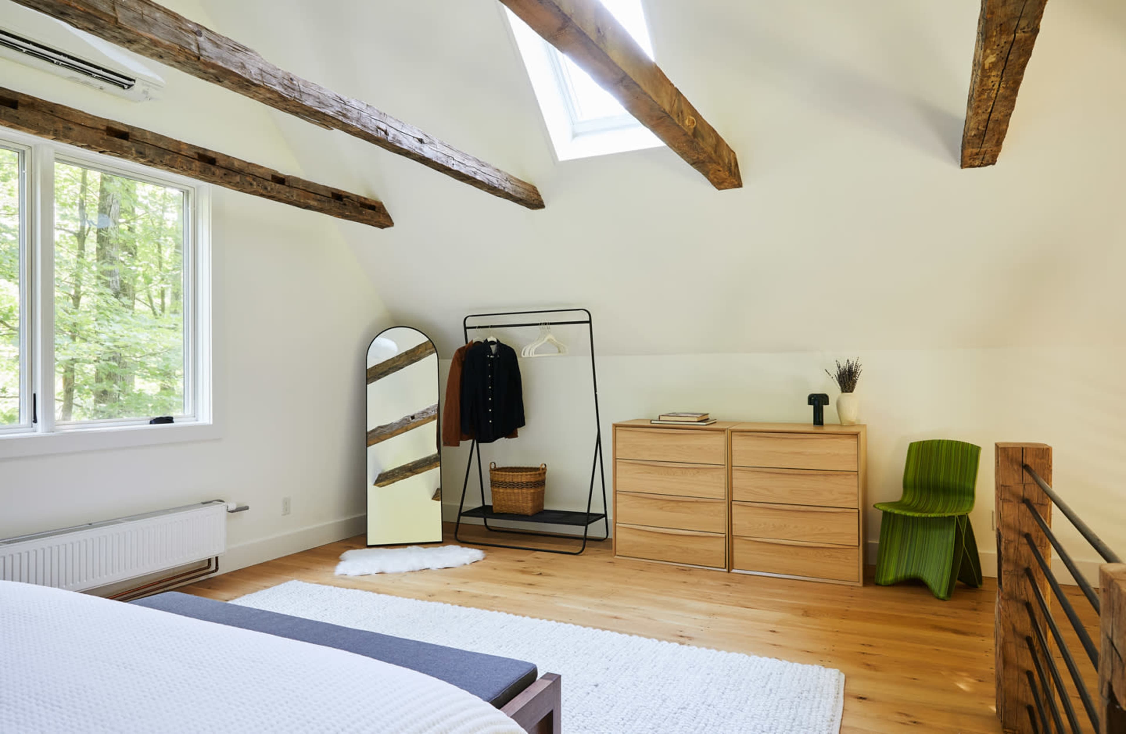 The bedroom features a sloped ceiling with exposed wooden beams, a large window, a minimalist wardrobe, a wooden chest of drawers, and a distinct green chair.