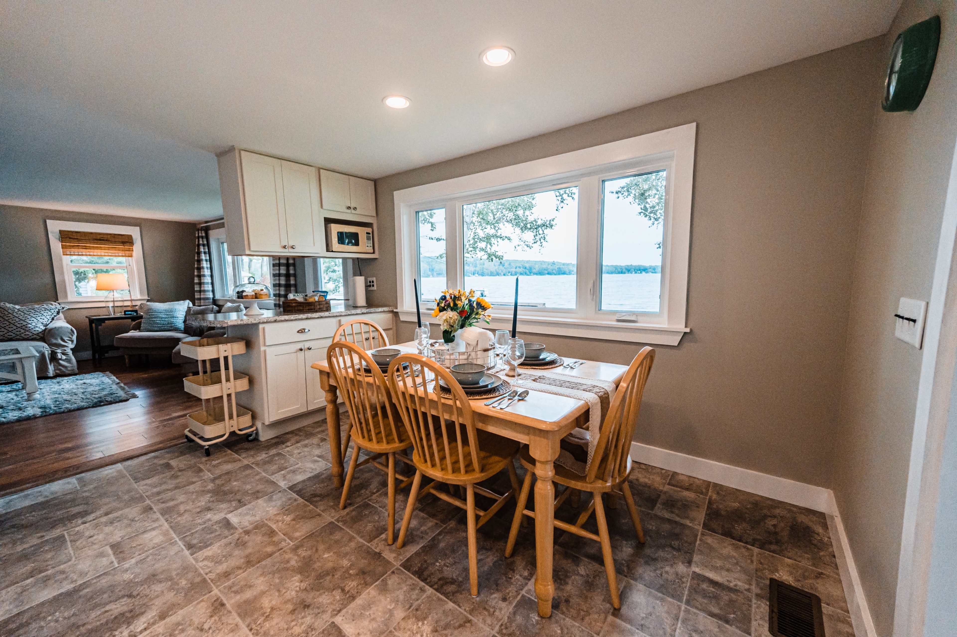 A dining area with a wooden table and chairs is set up near a large window overlooking a lake, with neutral-colored walls and tiled flooring.