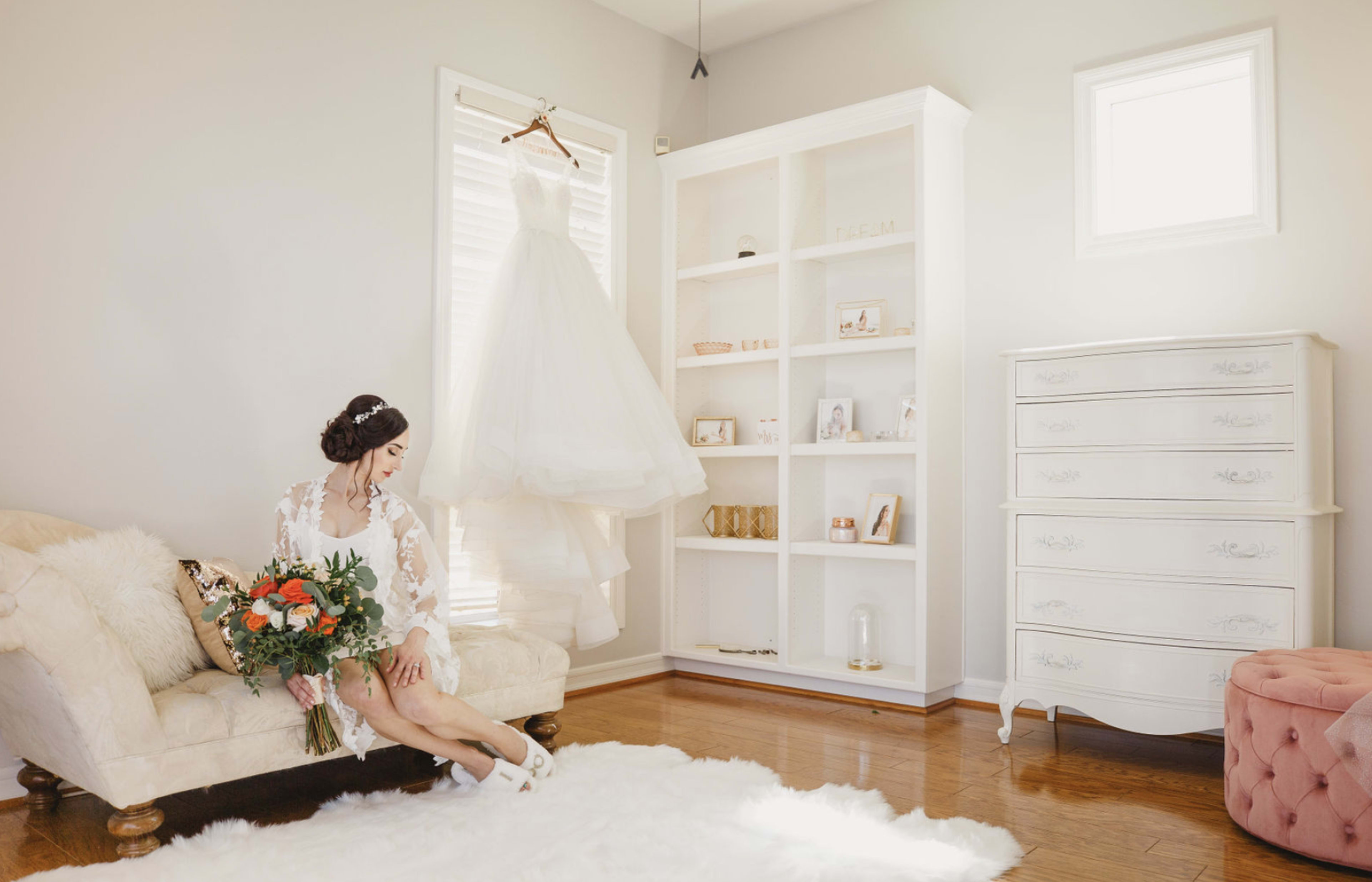 A bride in a white dress sits on a couch holding a bouquet of flowers, while her wedding gown hangs on a rack in a well-lit room.