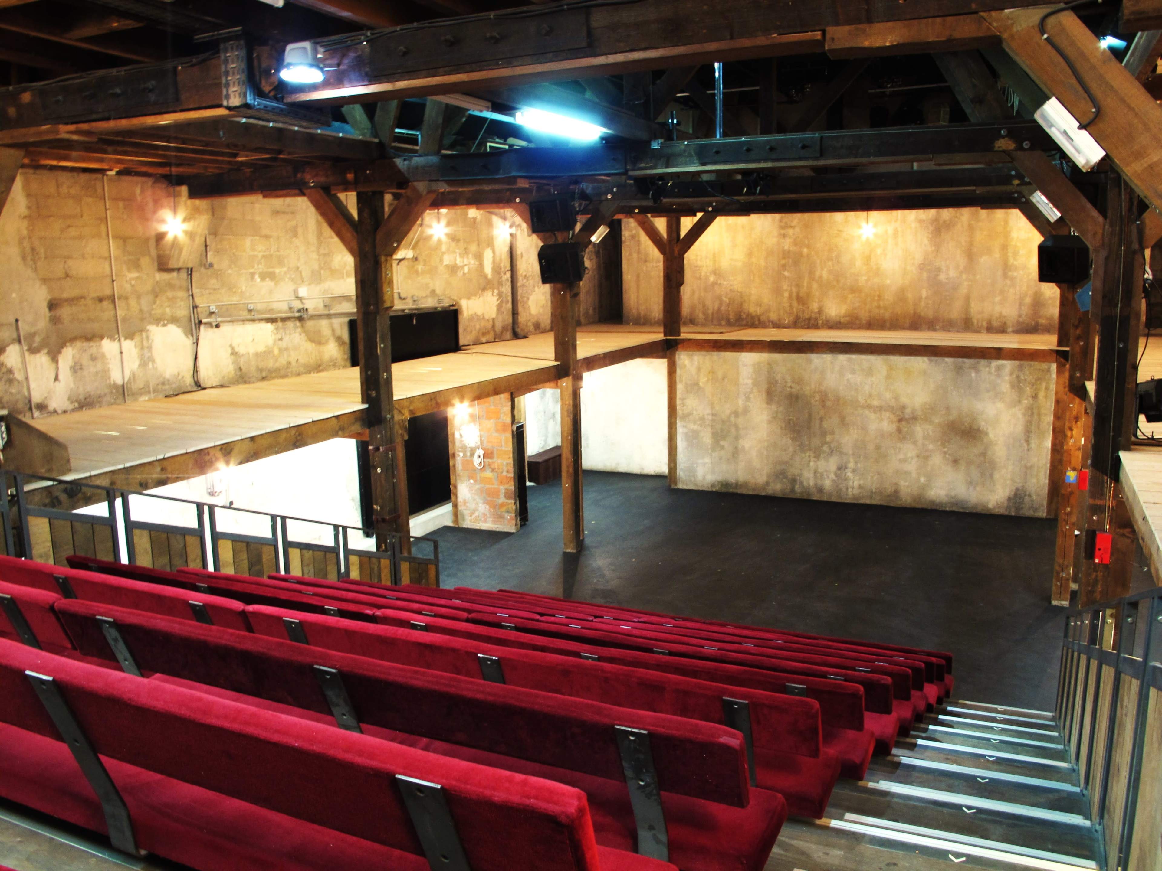 A spacious, empty theater with wooden beams and red seating arranged in tiers facing a simple stage area.