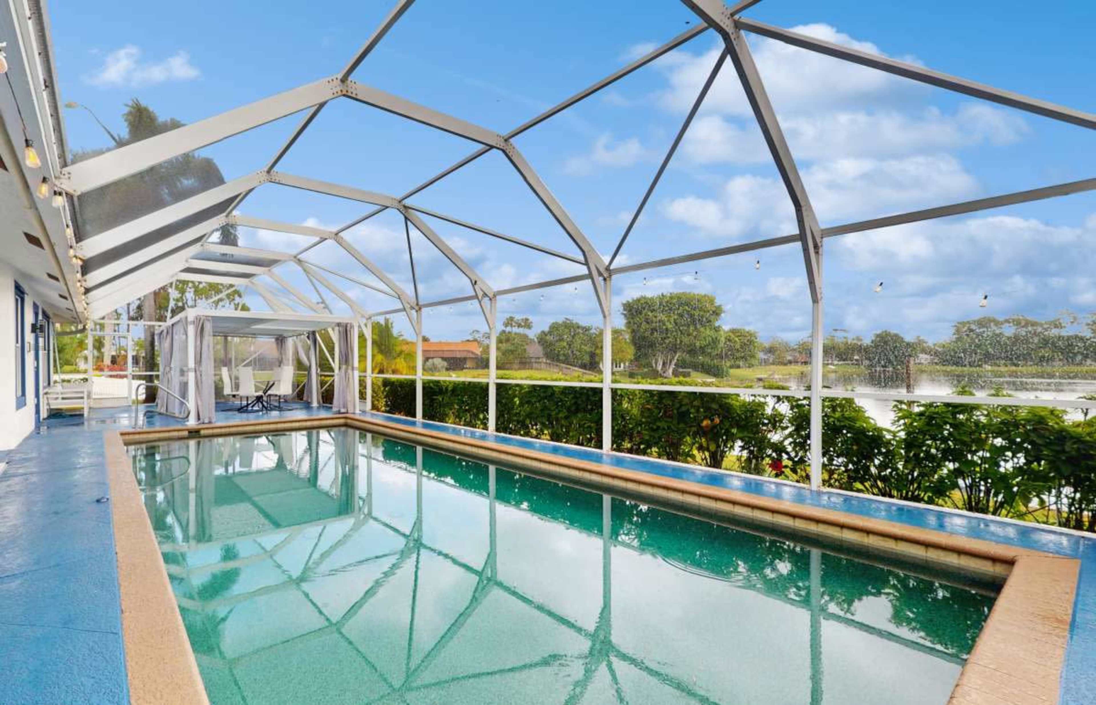 The image shows a screened-in swimming pool area surrounded by greenery and a clear blue sky.