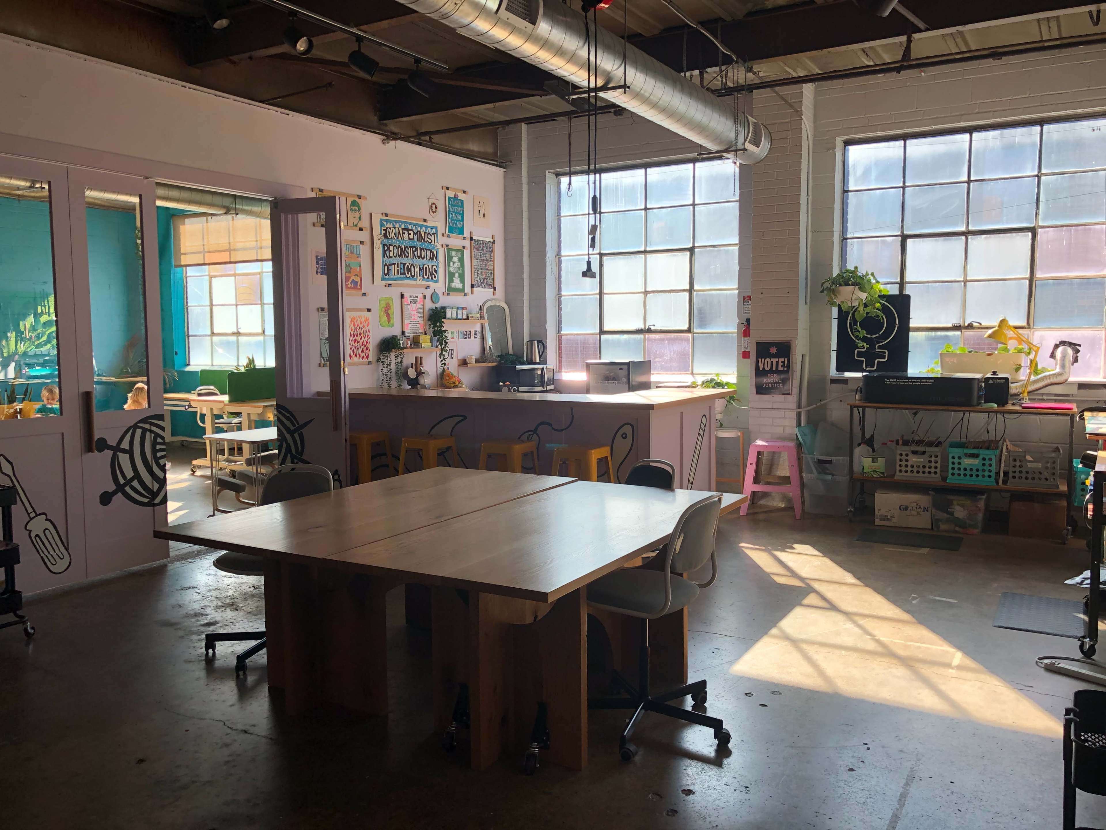 A well-lit communal workspace features two wooden tables, chairs, and colorful decor on the walls.
