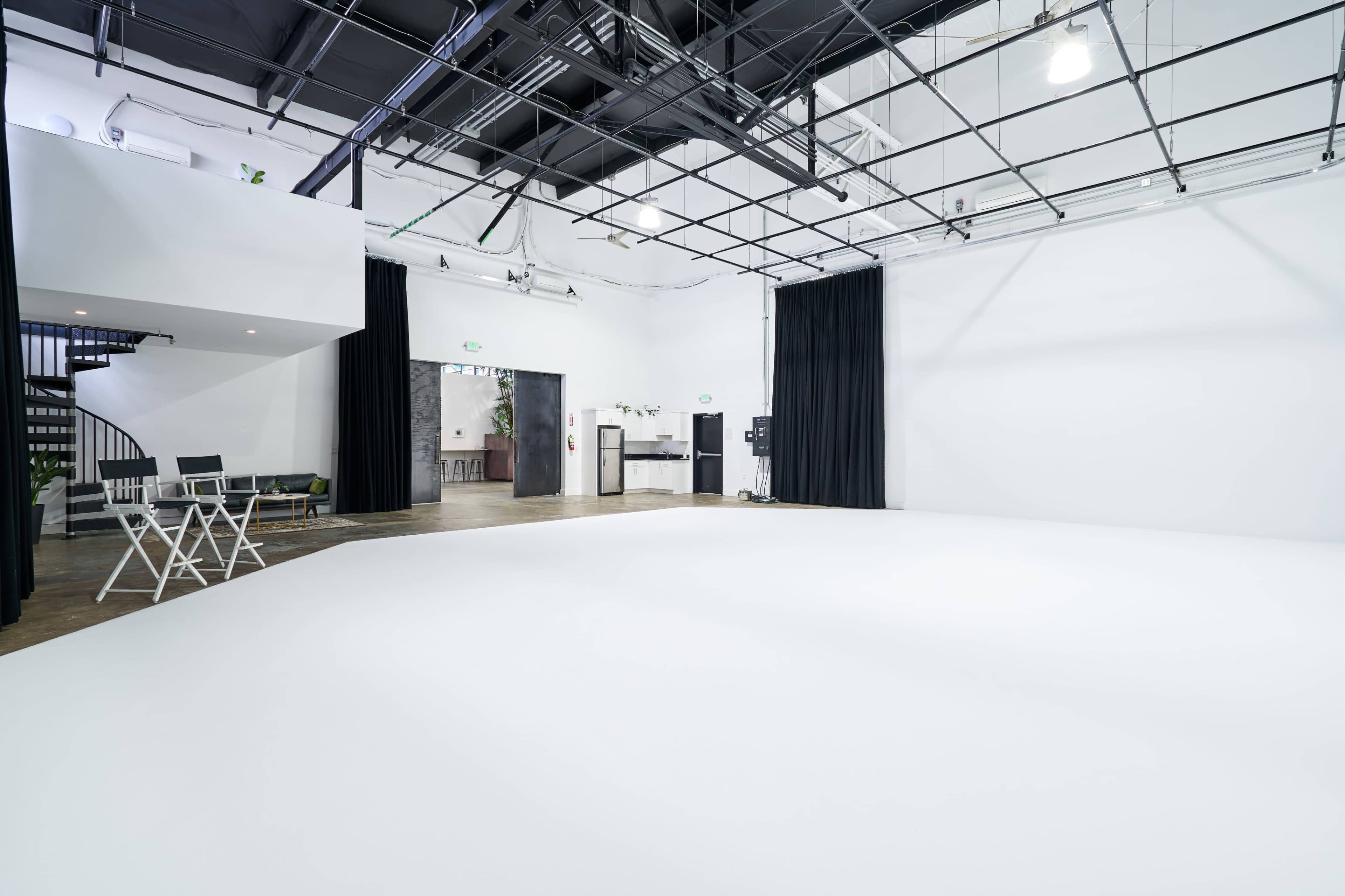 A spacious, empty studio features a white floor, black curtains, and a spiral staircase leading to an upper level.