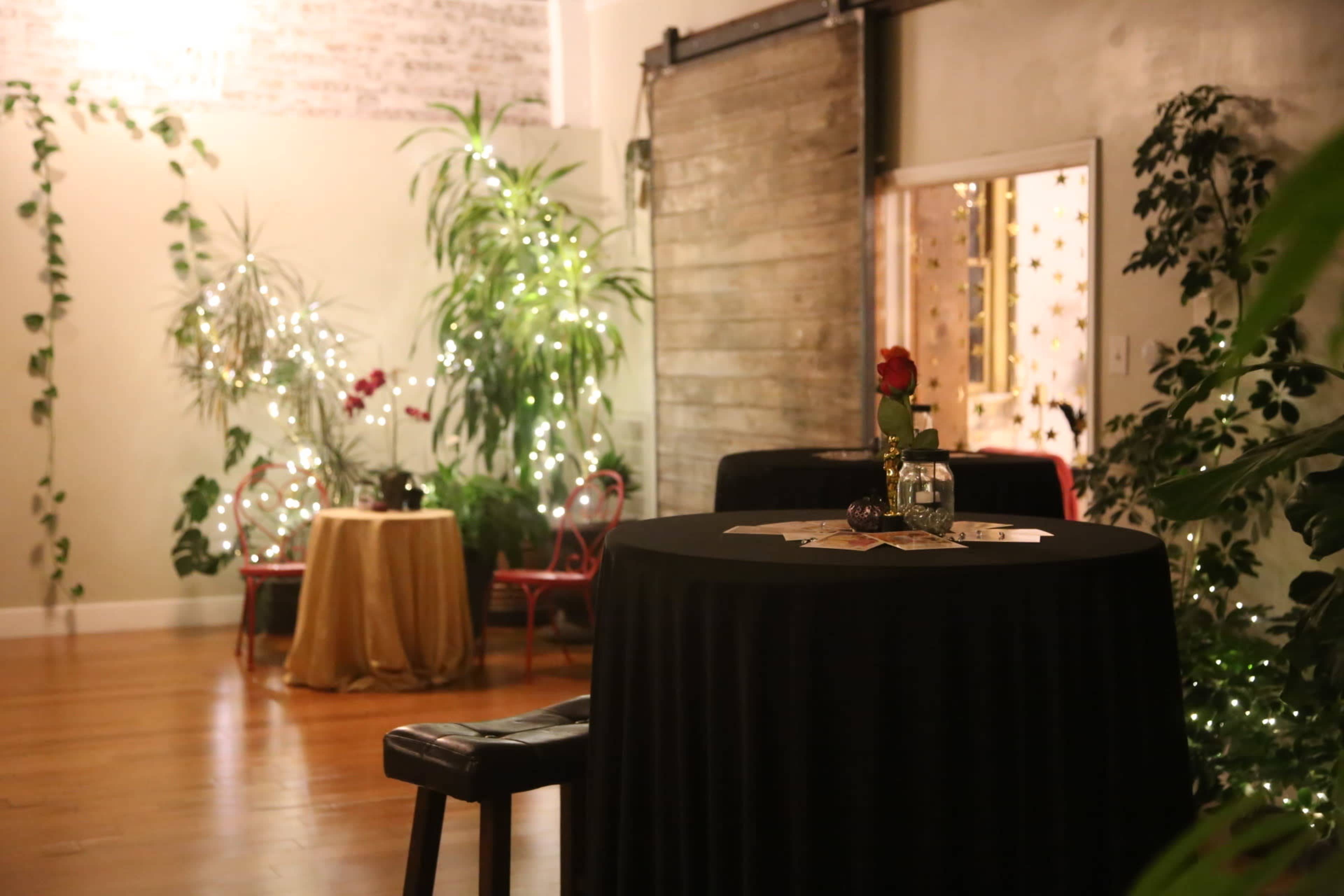 An indoor space decorated with plants and sparkling lights, featuring a black table set with a flower and surrounded by two chairs.