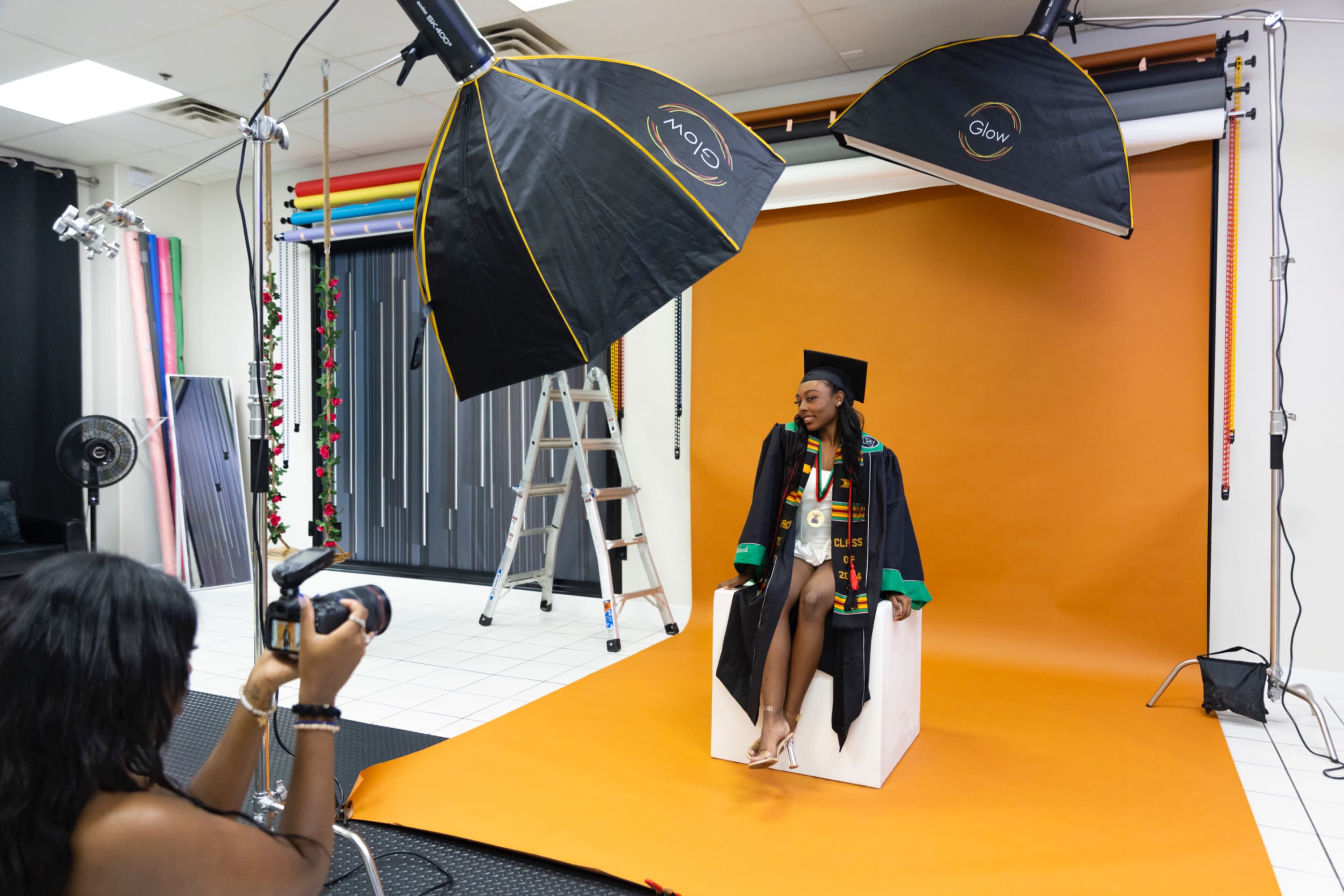 A graduate in a cap and gown poses on a stool in front of an orange backdrop while a photographer captures the moment.