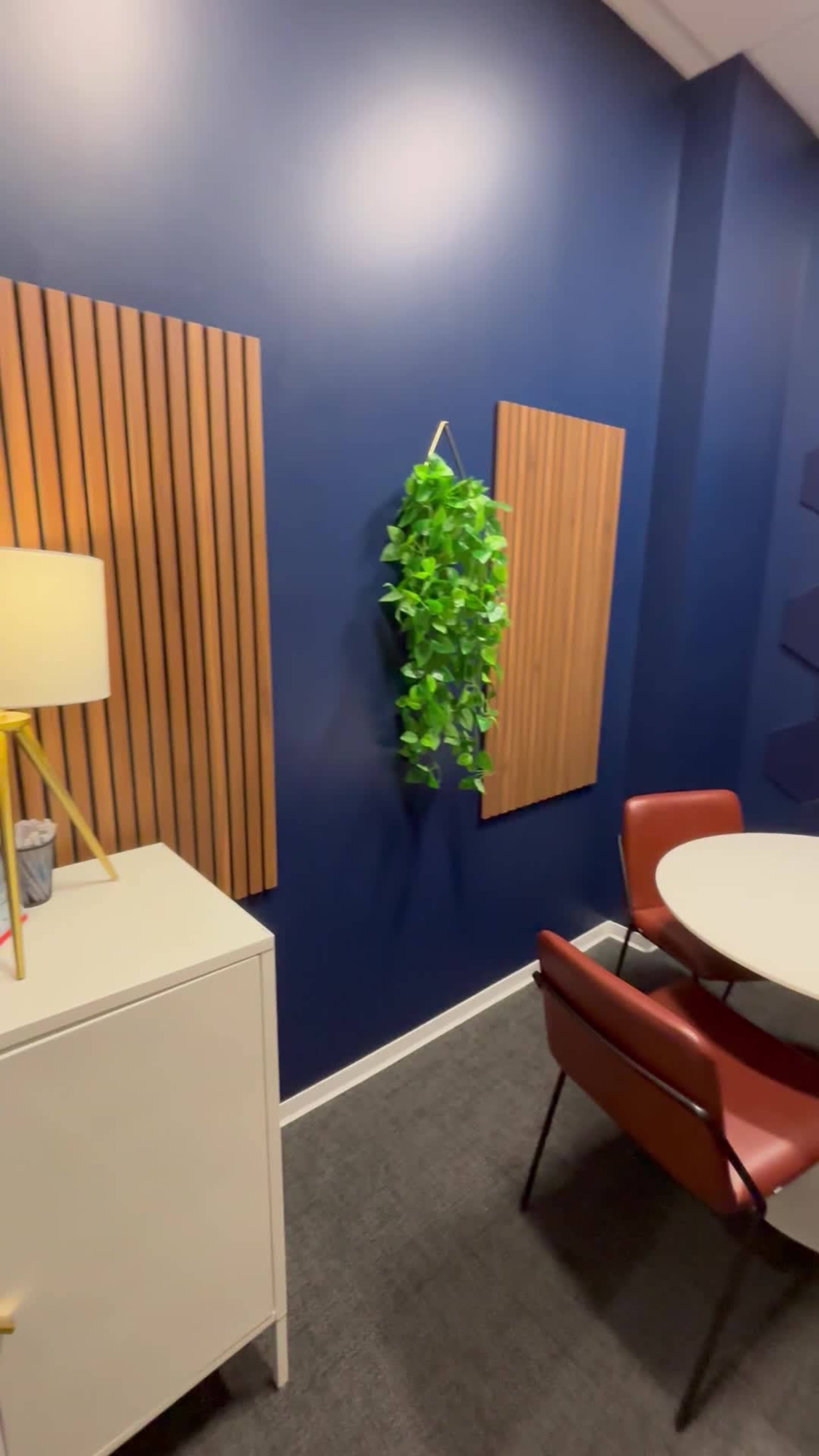 The image shows a small meeting room featuring a round table surrounded by chairs, a cabinet, wall panels, and a hanging plant against a dark blue wall.