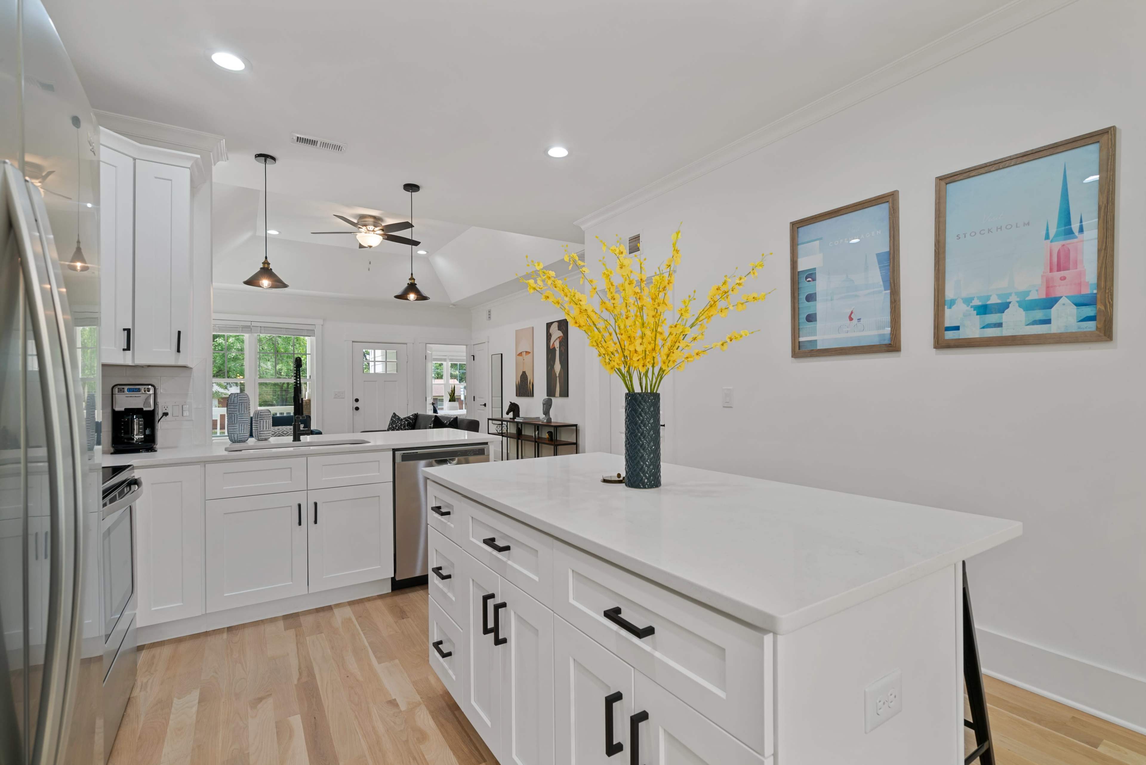 A bright kitchen features a central island with a vase of yellow flowers, surrounded by modern appliances and light-colored cabinetry.
