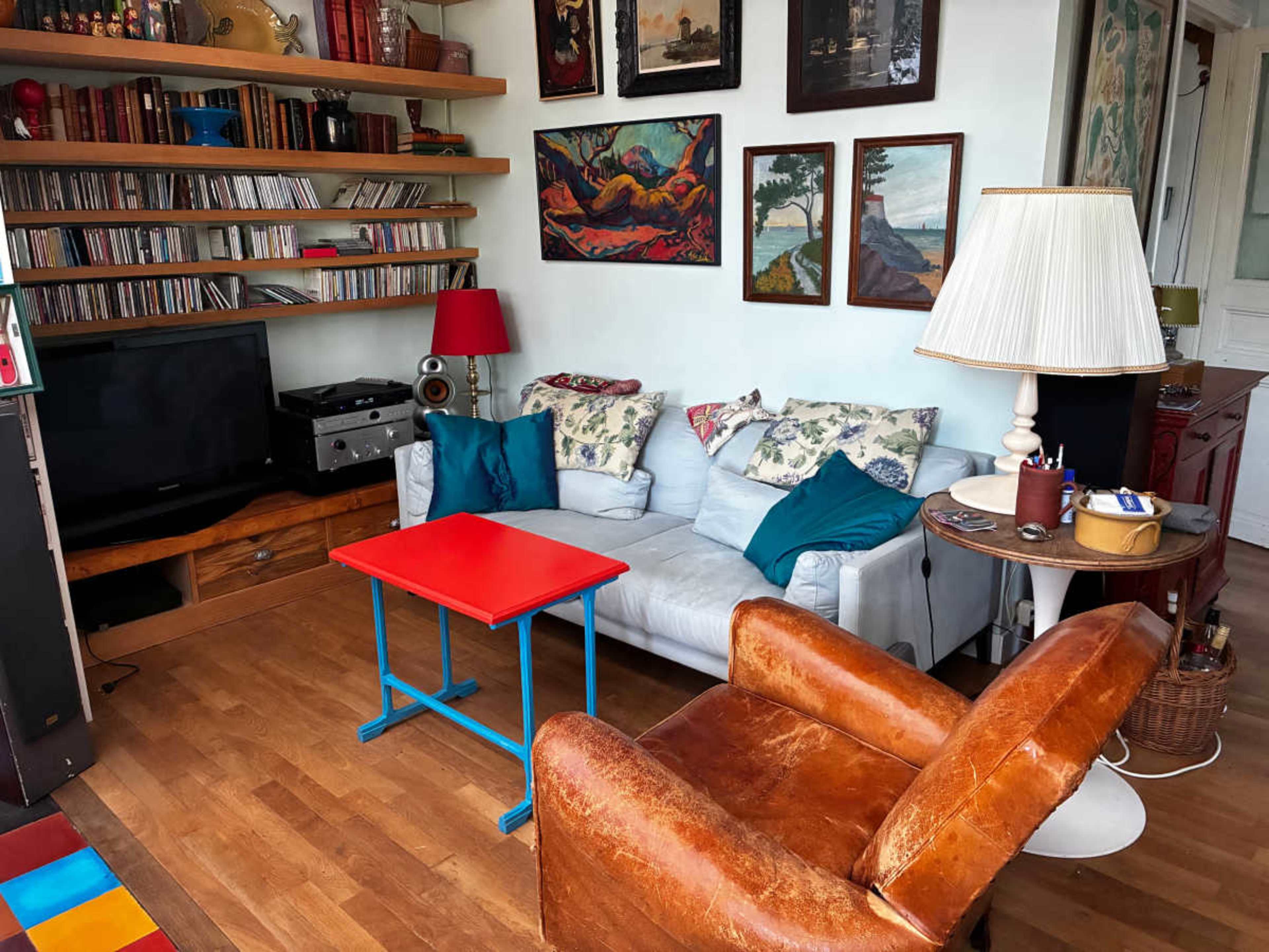 The image shows a cozy living room with a white sofa, orange armchair, red coffee table, and various framed artworks on the walls.