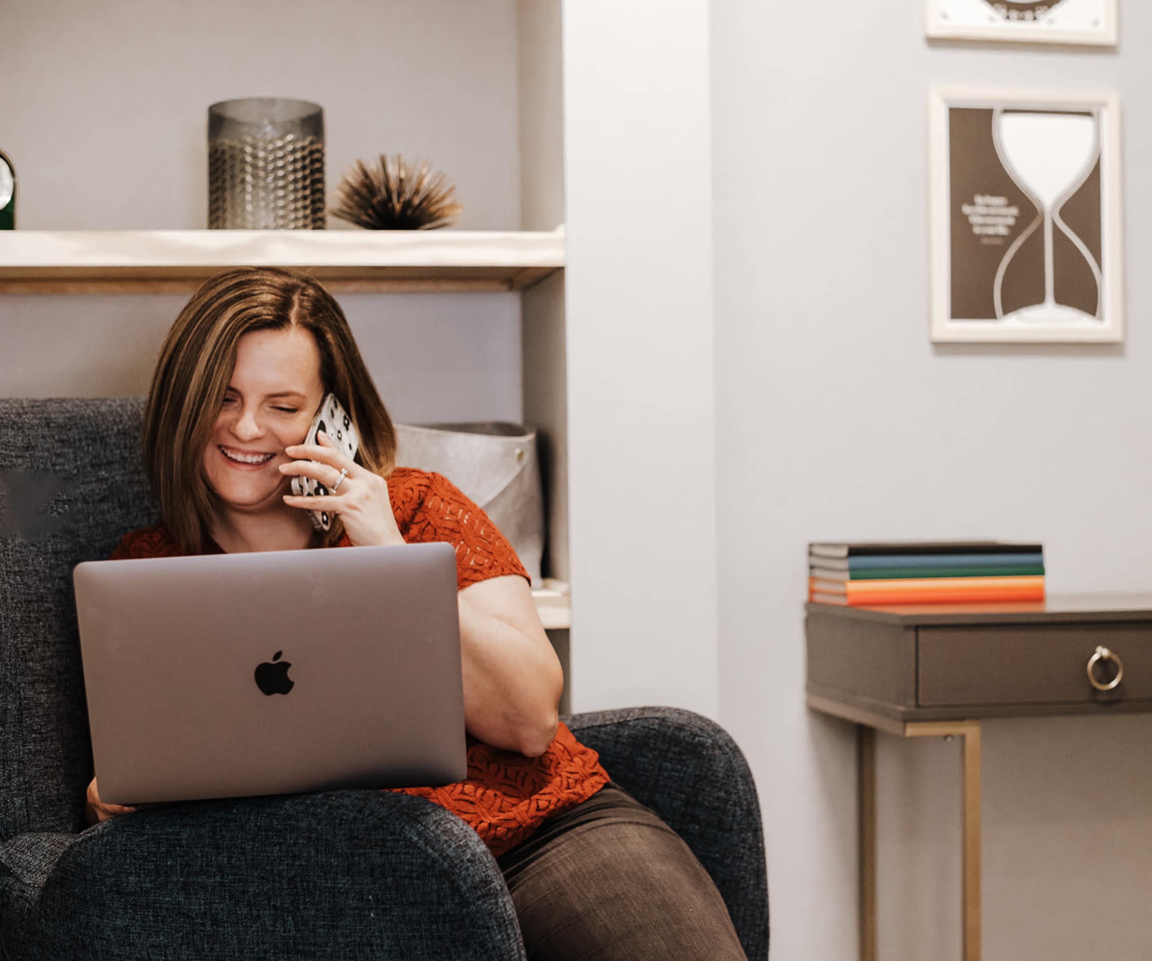 A woman sits on a blue chair using a laptop and talking on the phone in a neatly arranged room with shelves and a side table.