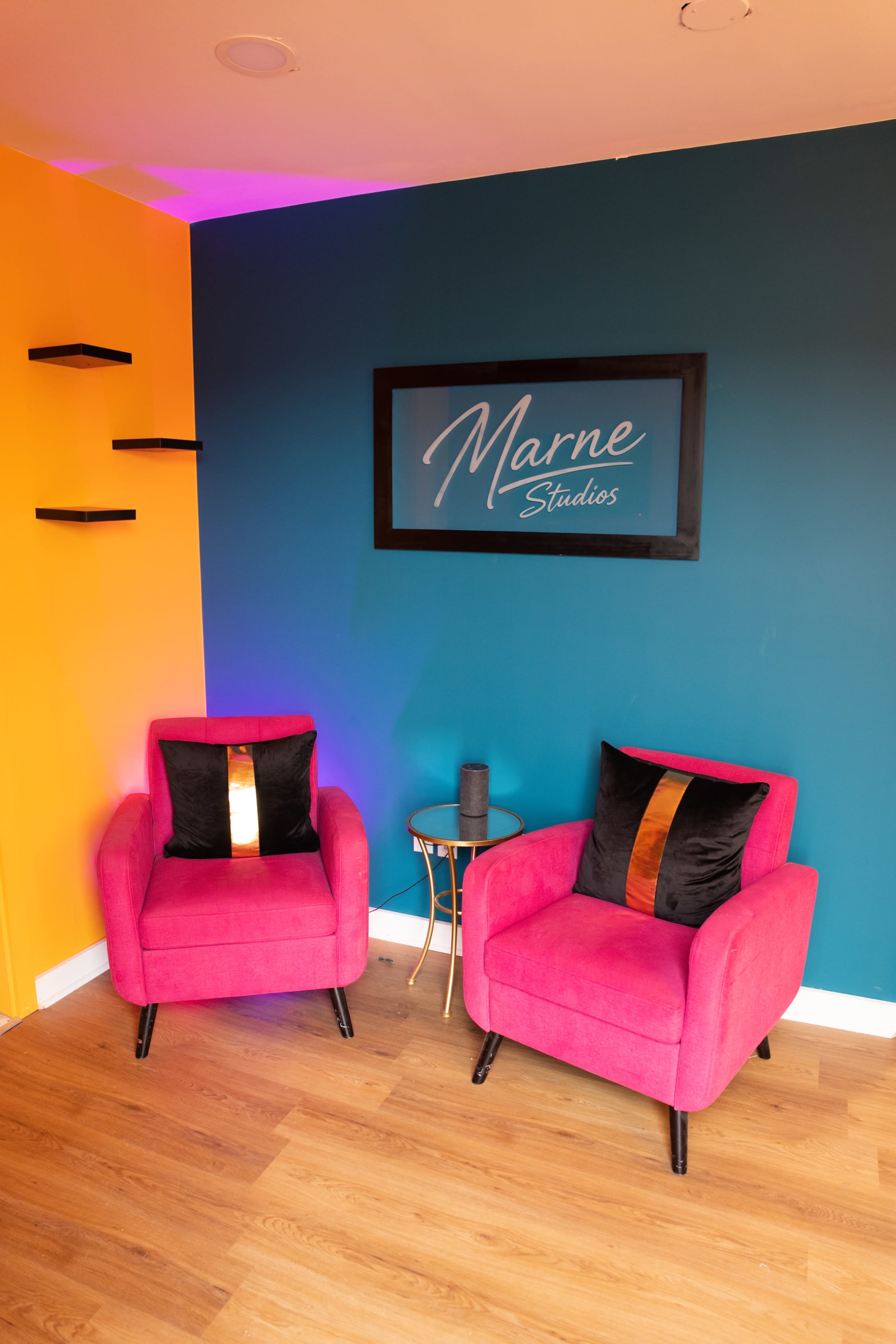 The image shows a cozy waiting area with two pink armchairs and a small side table against a teal and orange accent wall, featuring a sign that reads "Marne Studios."