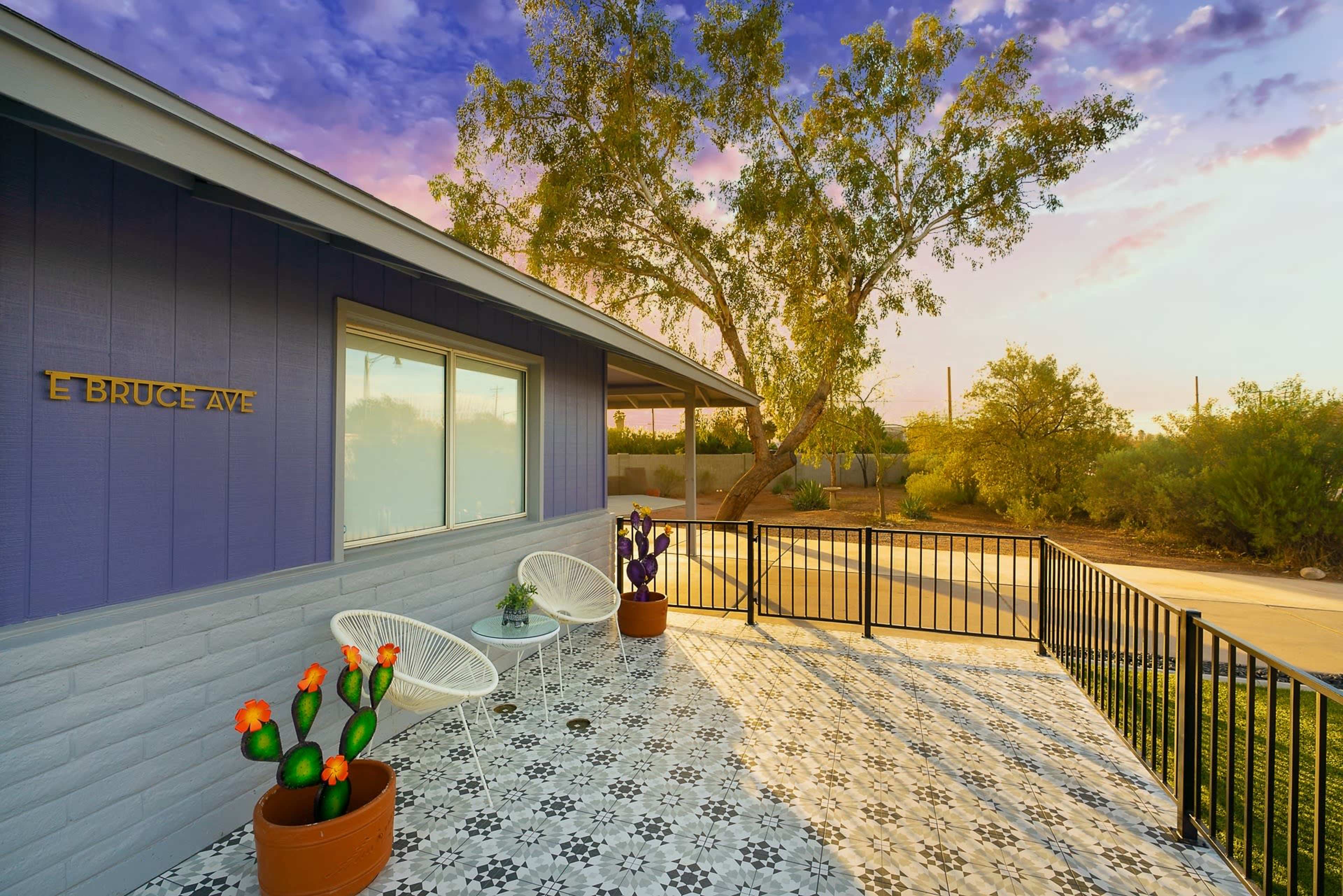 A colorful patio with white chairs, potted cacti, and decorative plants, alongside a modern home under a sunset sky.