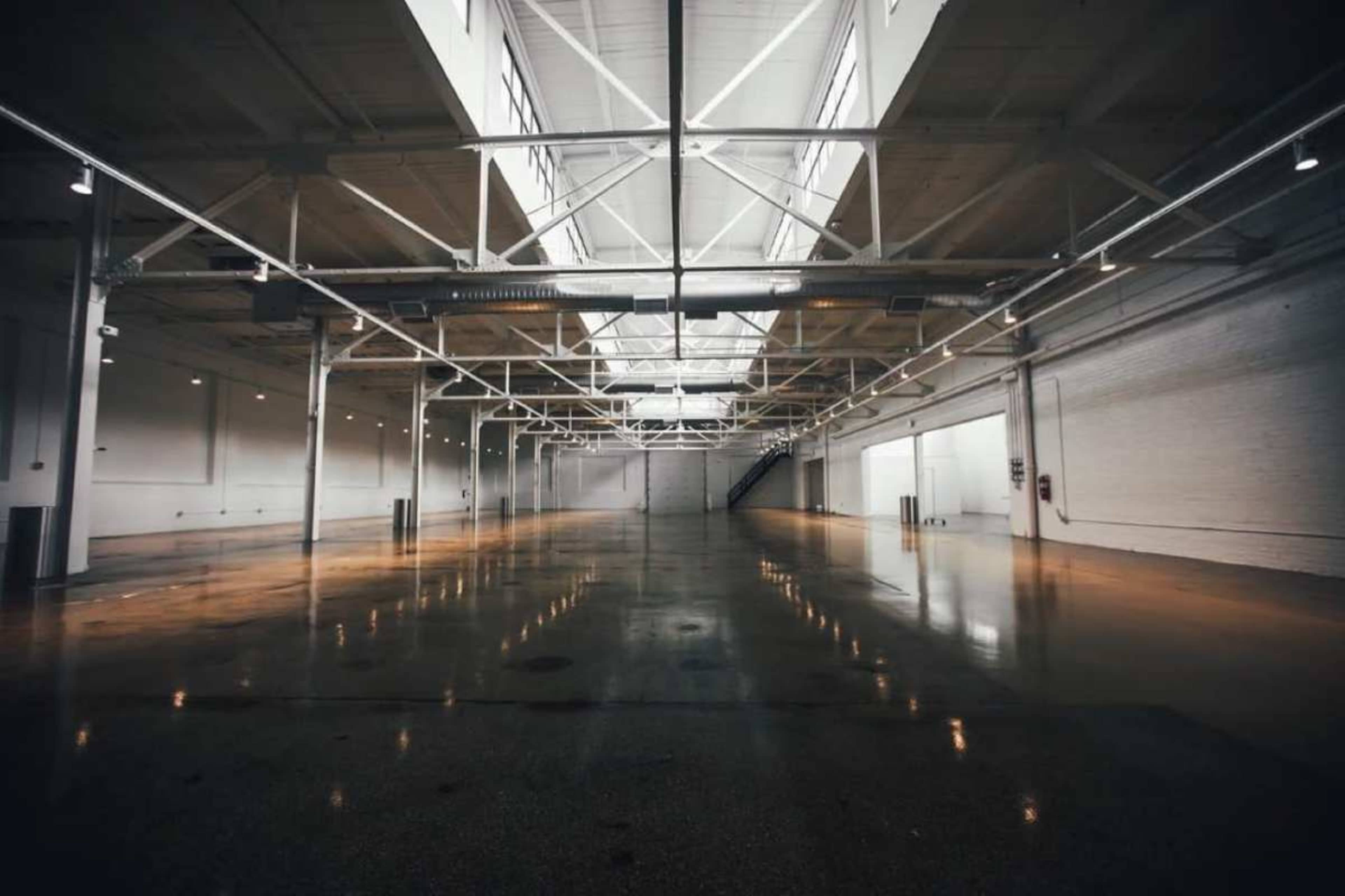 The image shows a large, empty industrial space with high ceilings, exposed beams, and polished concrete floors.