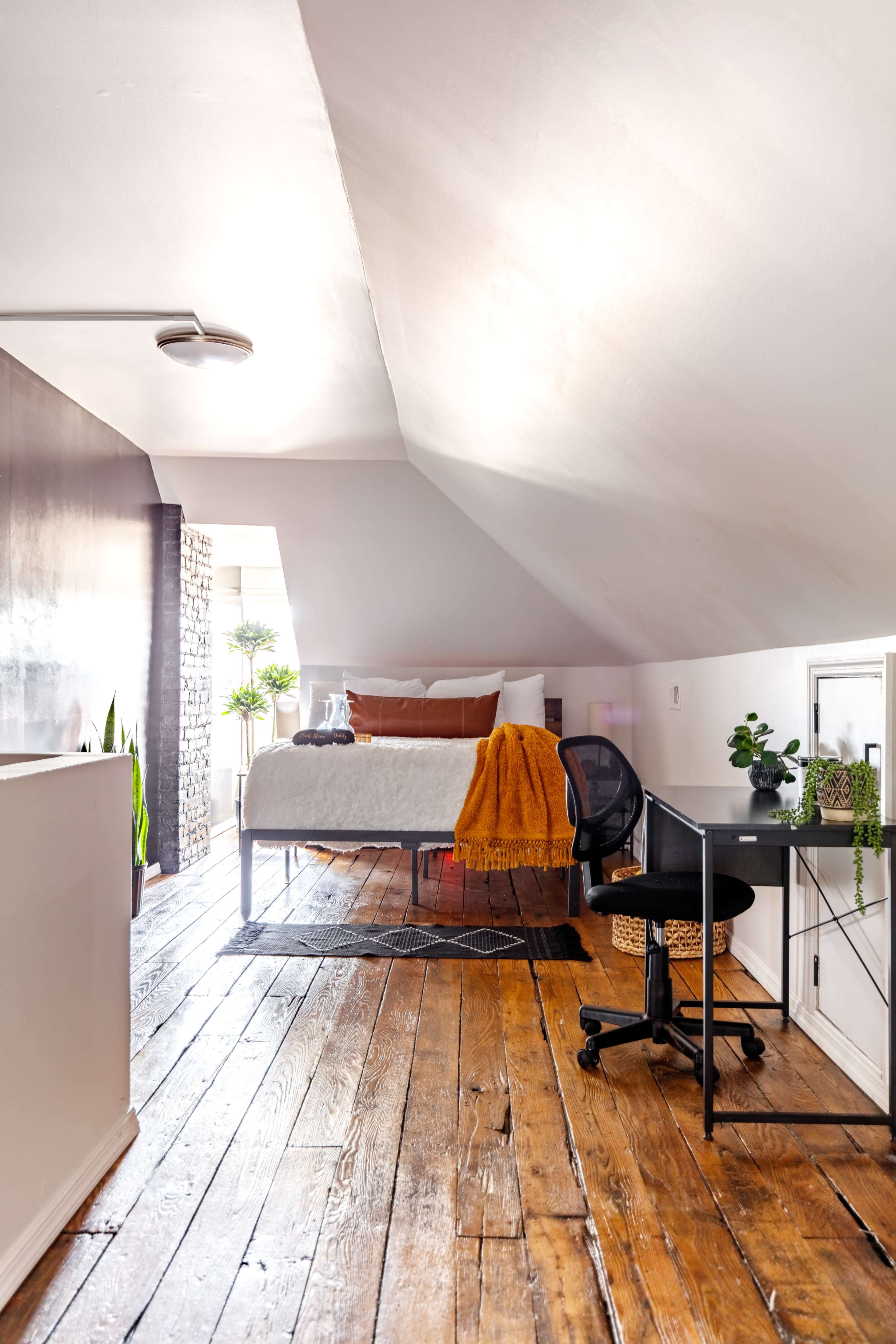 The image shows a bright attic room featuring a bed, a desk with a chair, and wooden floors, with plants arranged around the space.