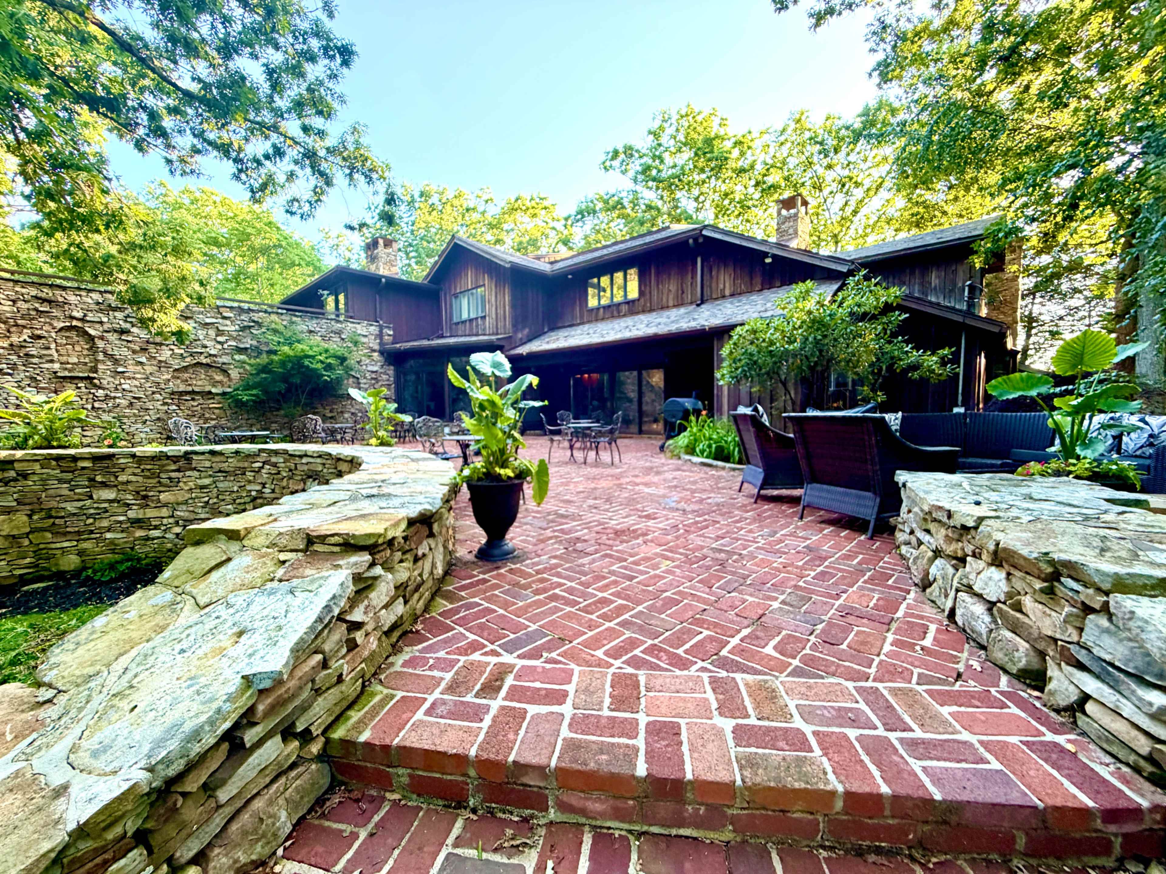 The image shows a large wooden house with a brick pathway leading to a patio, surrounded by greenery and stone walls.