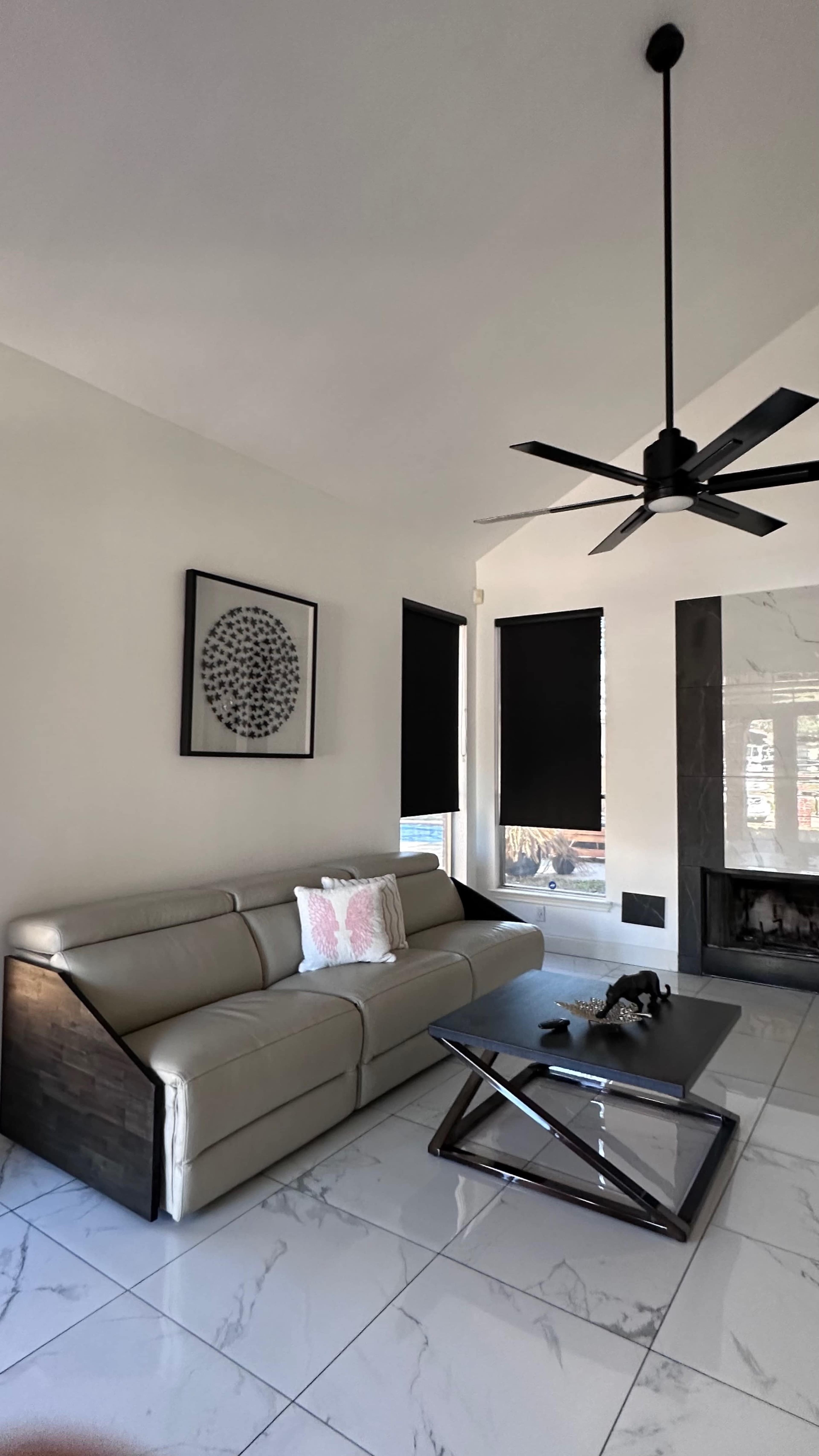 The scene features a modern living room with a light-colored leather sofa, a black coffee table, and a fireplace surrounded by large windows with black blinds.