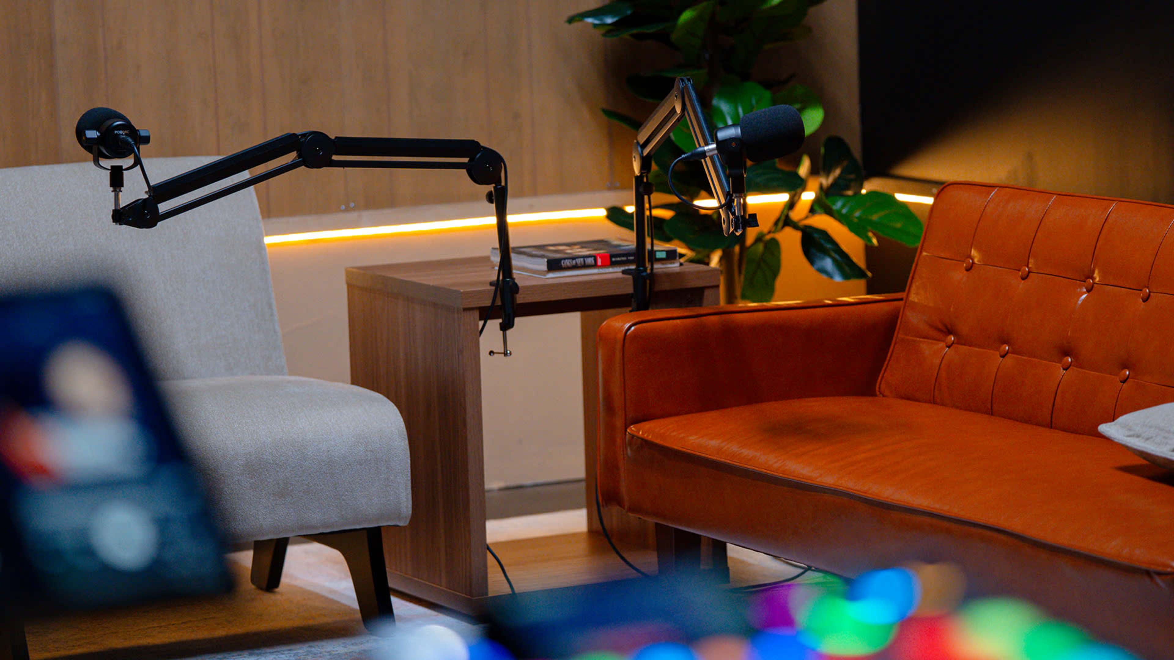 The image shows a cozy setup with an orange leather sofa and a light gray armchair, featuring two microphones mounted on adjustable arms positioned on a wooden side table in a well-lit room.