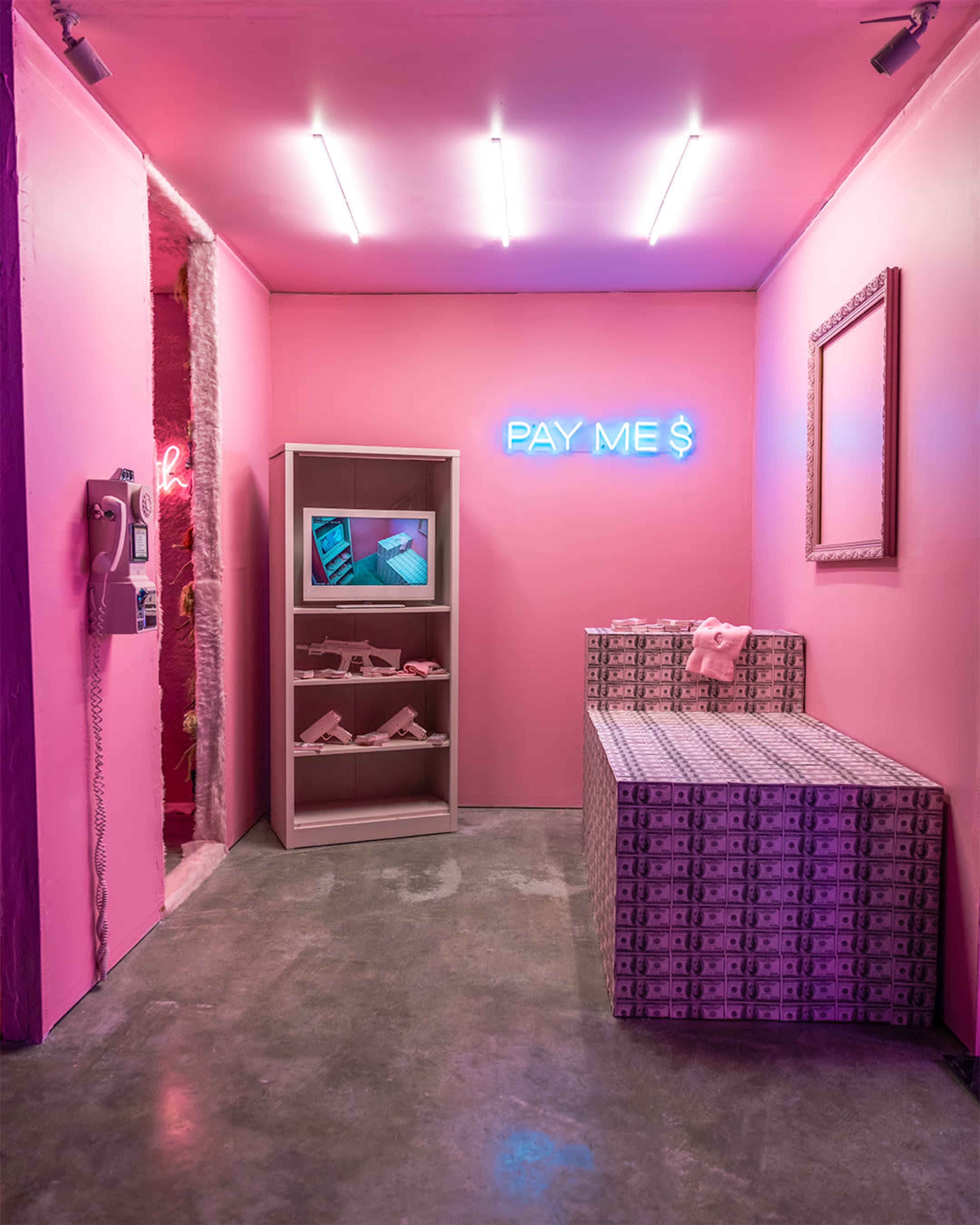 A pink-themed room featuring a television on a shelf, a neon sign reading "PAY ME $," and a couch covered in money-patterned fabric.