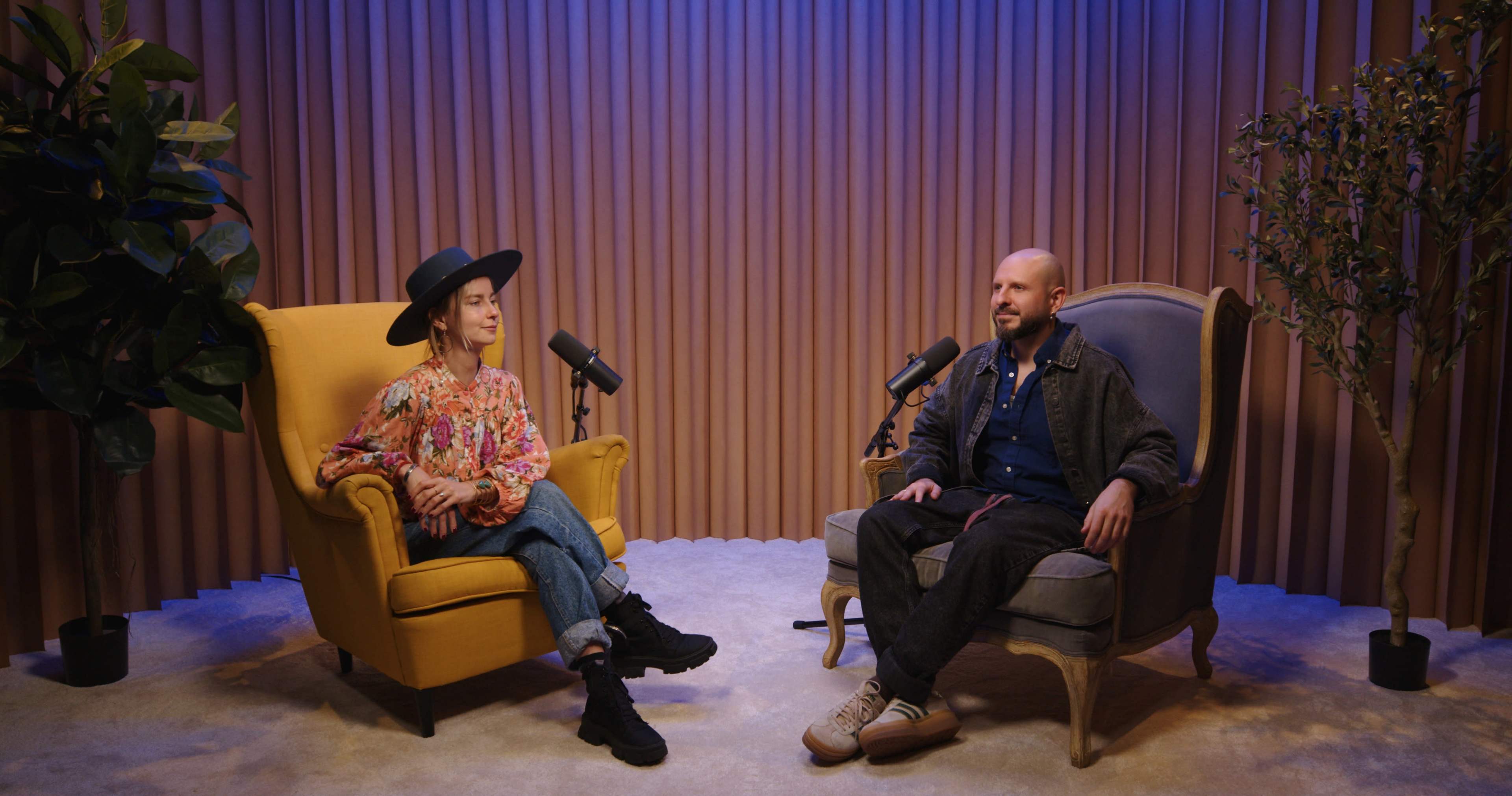 A woman in a floral blouse and hat sits on a yellow armchair, while a man in a jacket sits on a gray armchair, both positioned in a well-lit studio setting with microphones nearby.
