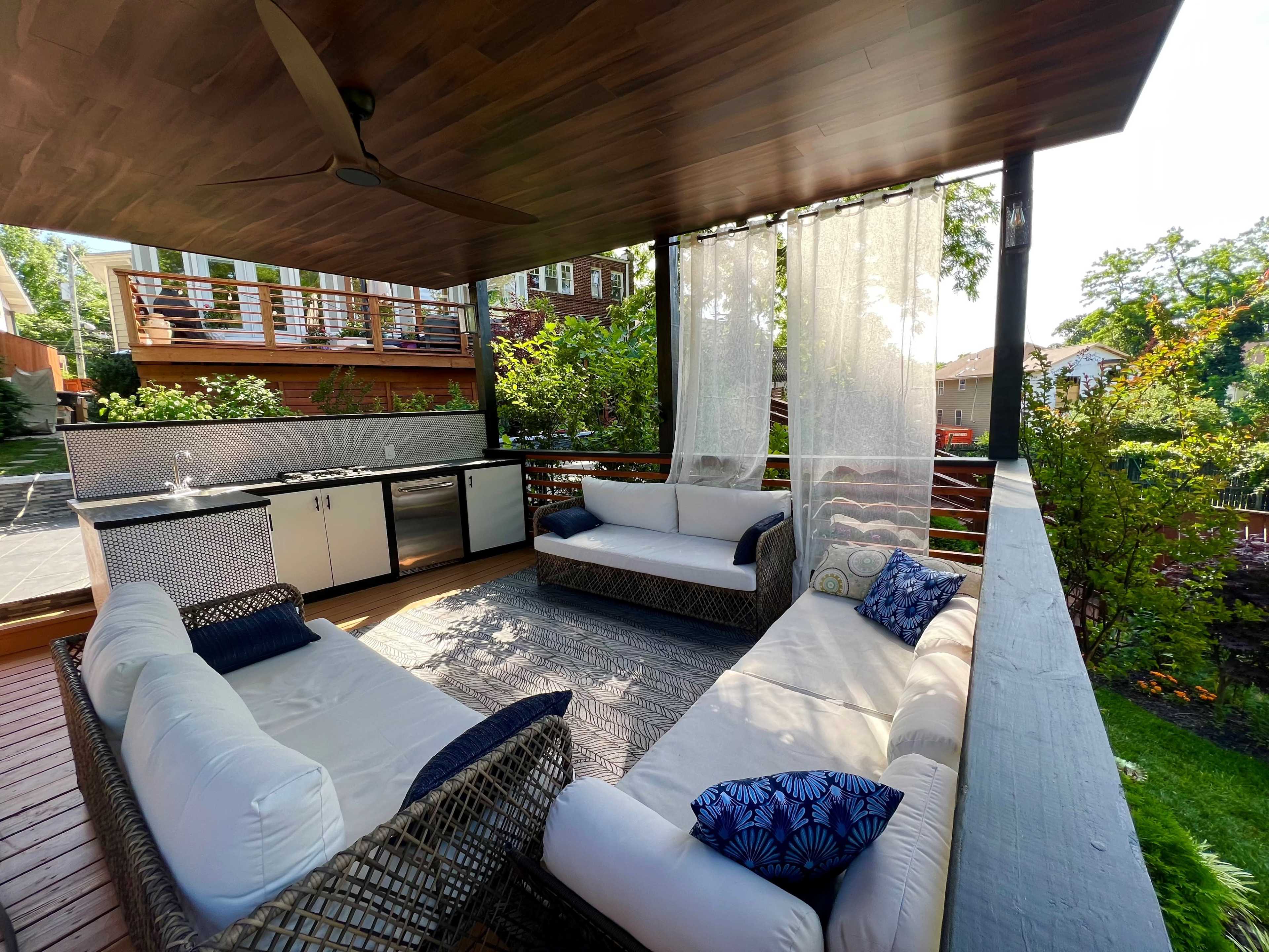 A covered outdoor seating area with a kitchen and comfortable sofas surrounded by greenery.