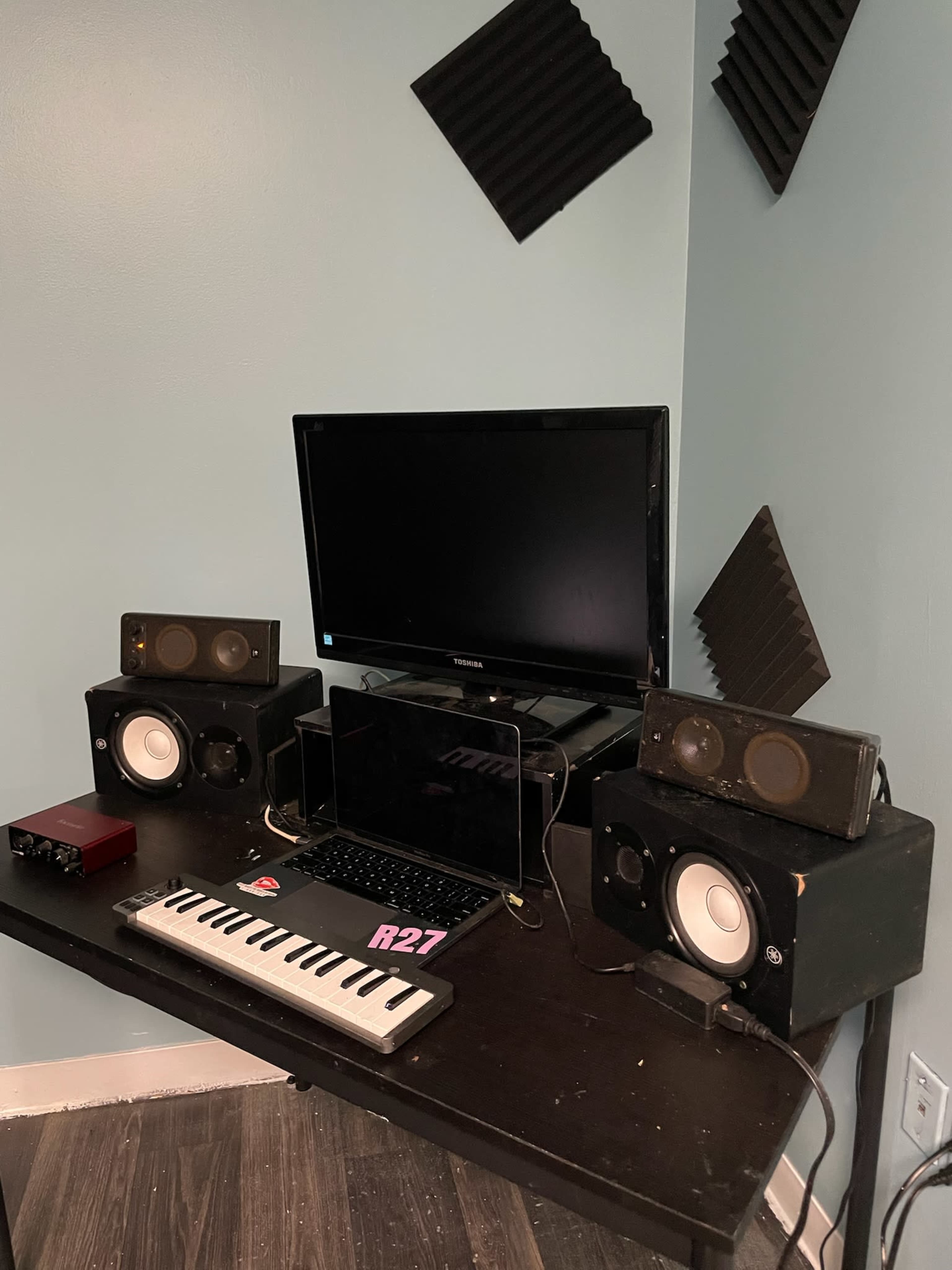 A desktop setup features a monitor, speakers, a keyboard, and an audio interface arranged on a dark table against a blue wall.