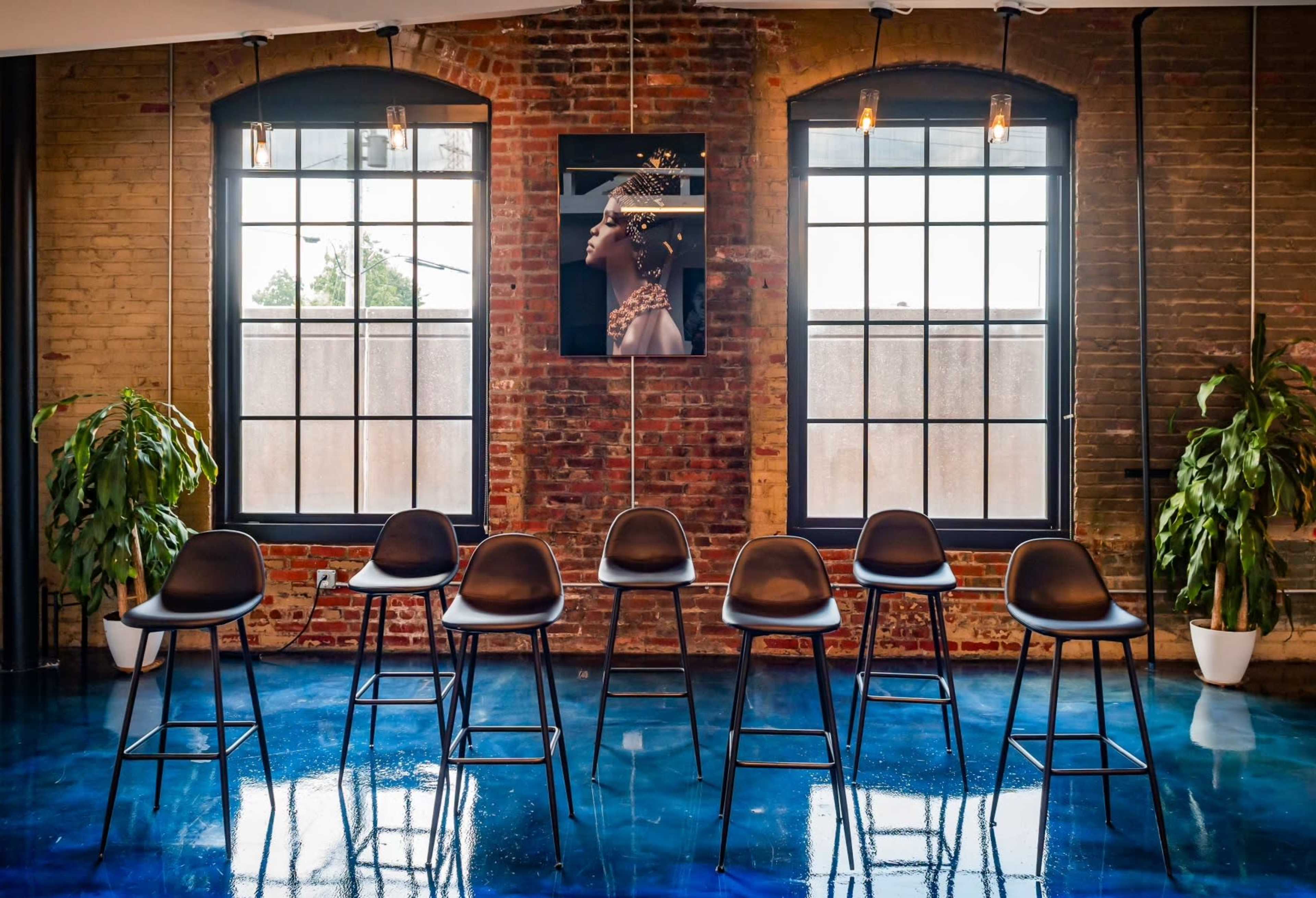 A line of black bar stools is arranged in front of large windows with a brick wall and a framed portrait hanging between them.
