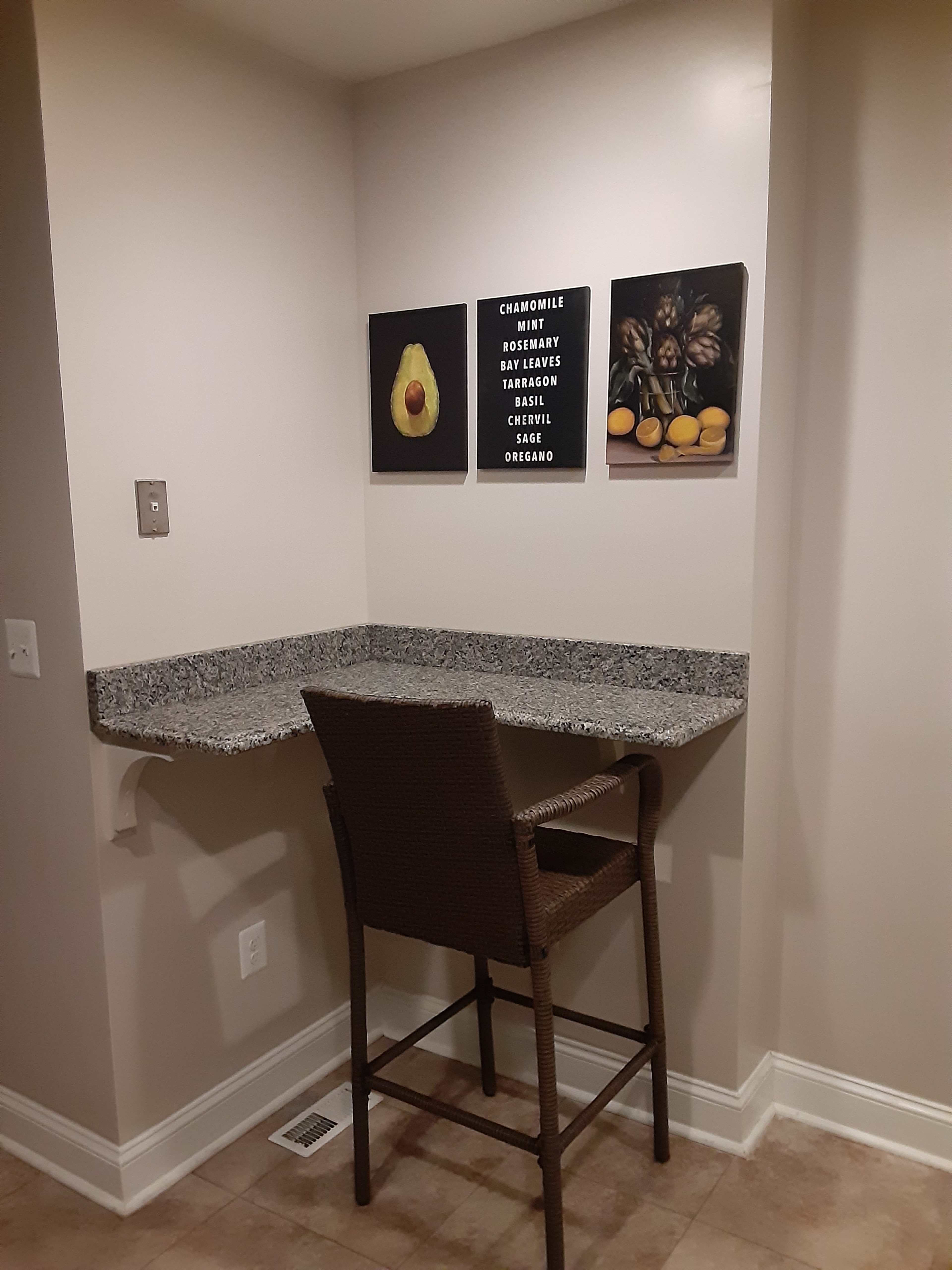 A corner nook features a granite countertop and a single barstool, with three framed images of vegetables and herbs hanging on the wall above.