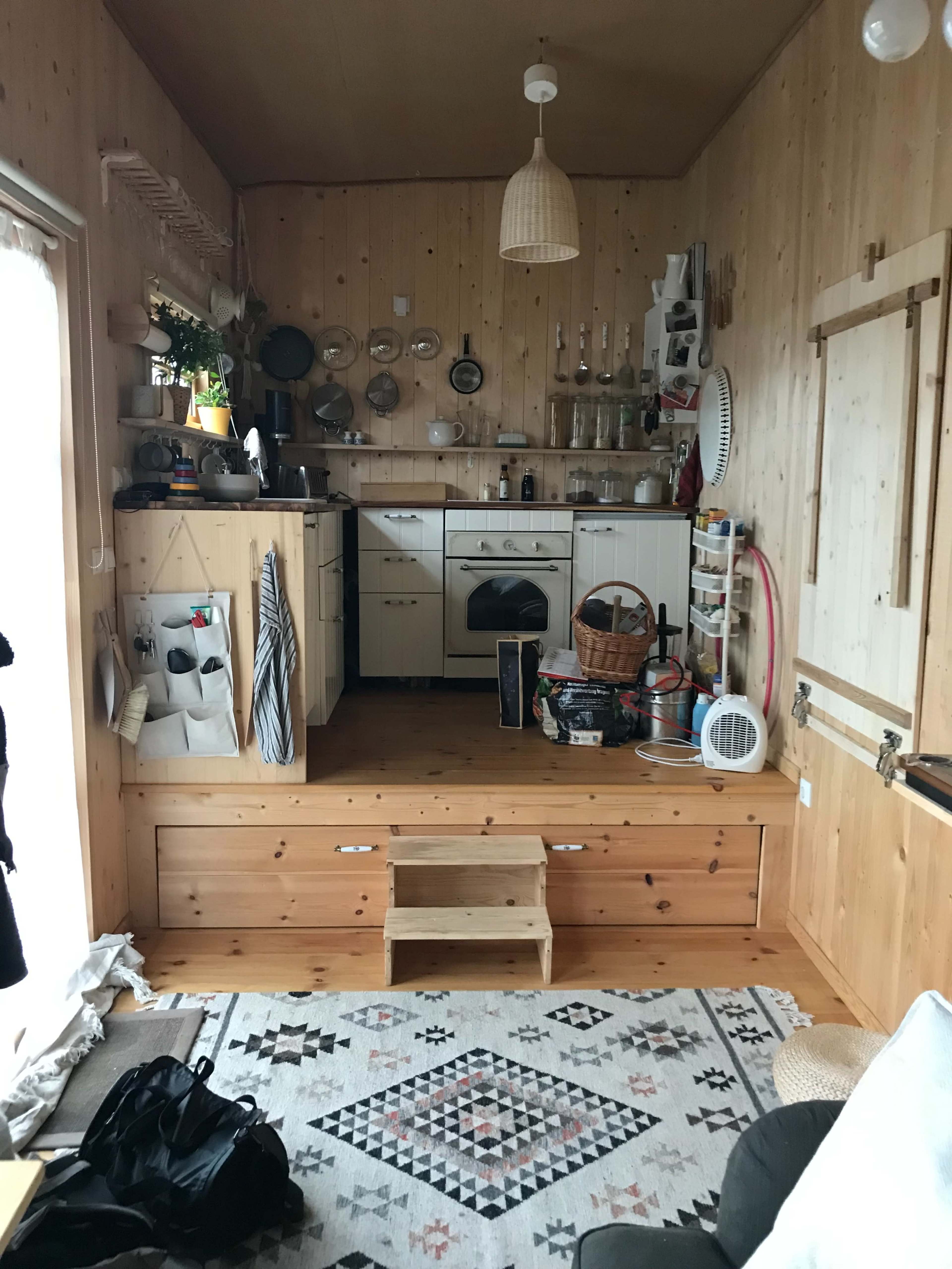 The image shows a wooden interior kitchen with counters, shelves, and various kitchen utensils neatly arranged, along with a small staircase leading to a raised platform.