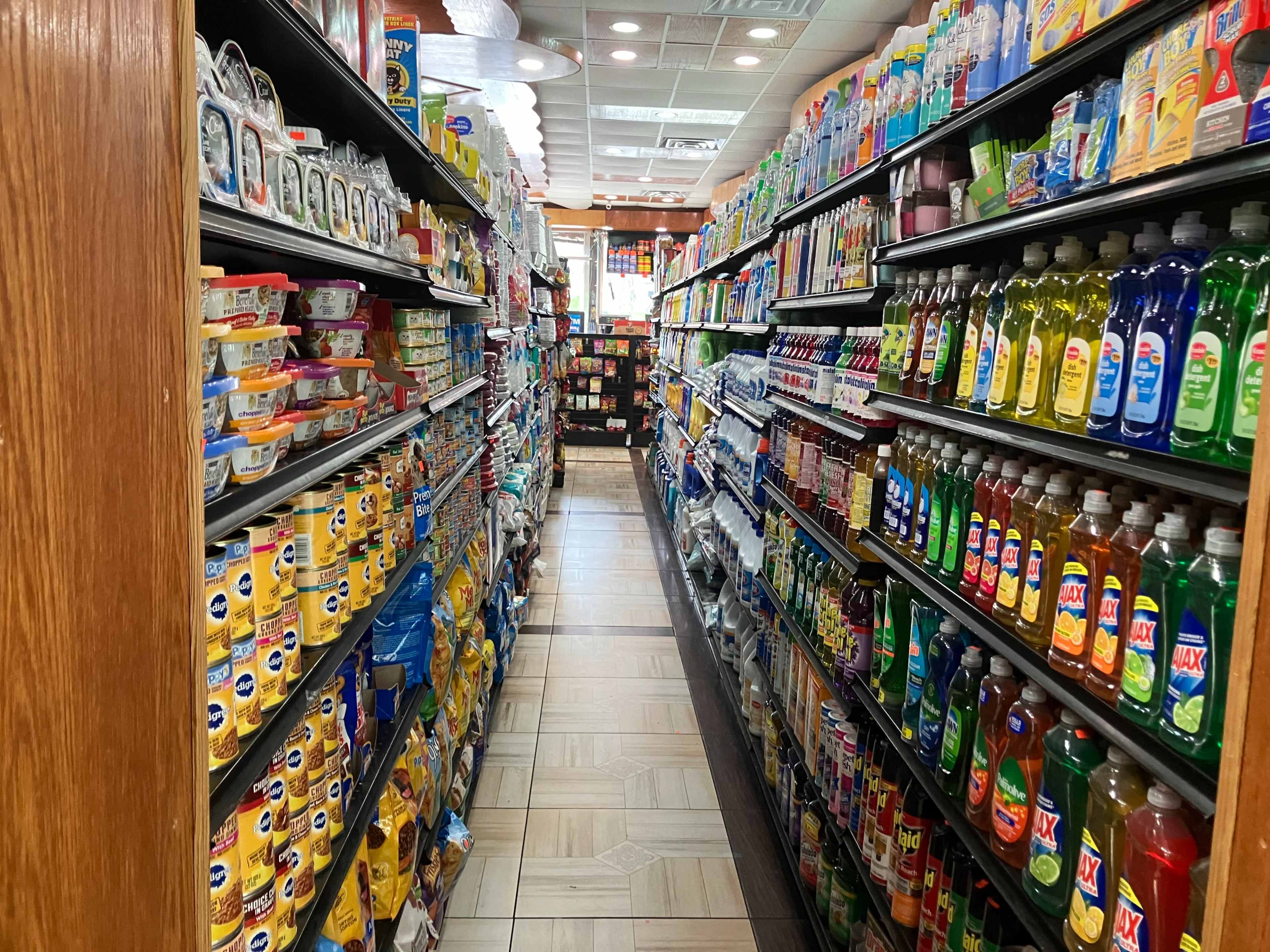 A narrow aisle in a grocery store lined with shelves stocked with various household cleaning products and food items.