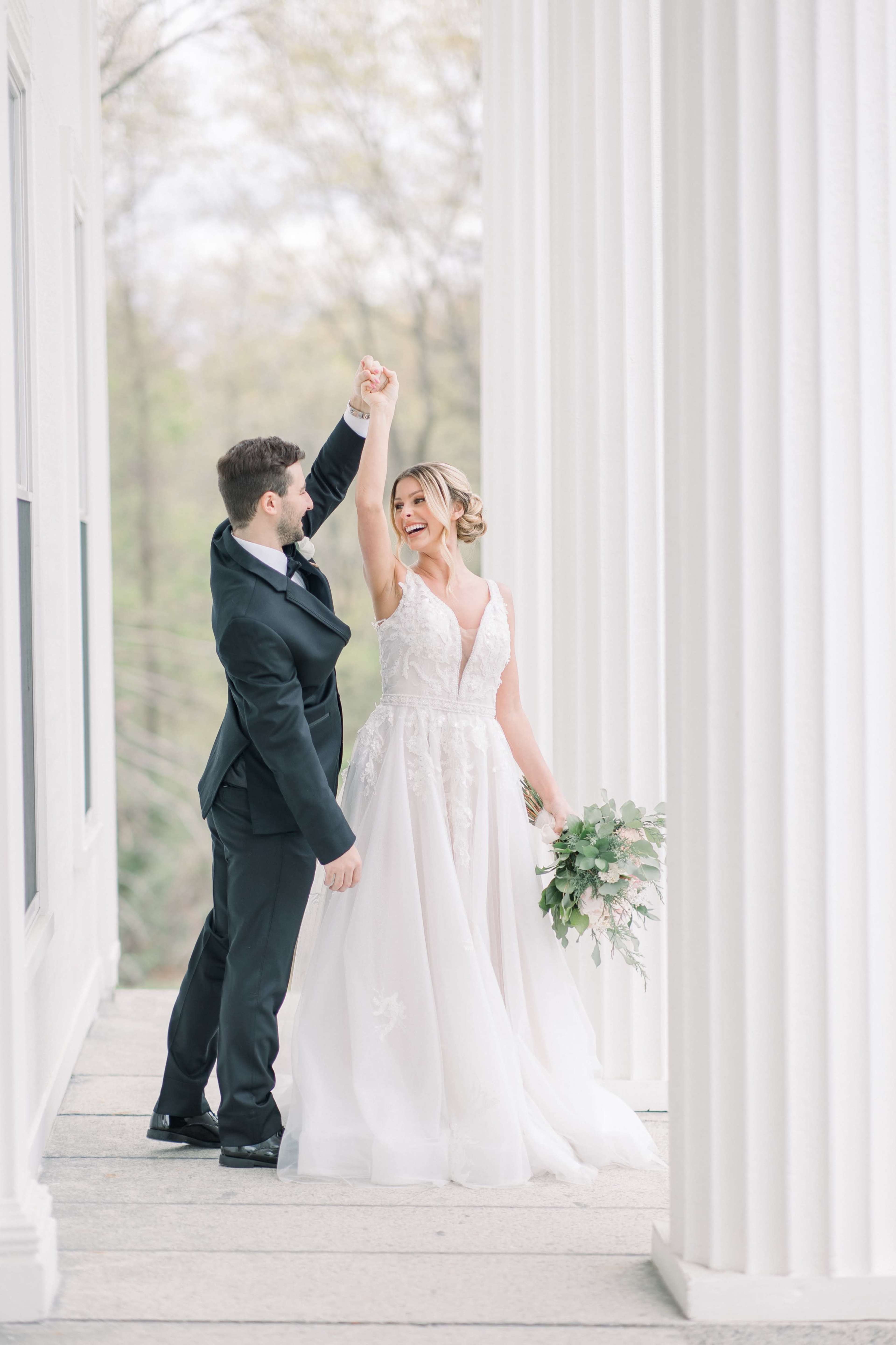 A bride in a flowing white dress holds hands with a groom in a black tuxedo, both smiling and dancing under a colonnade.