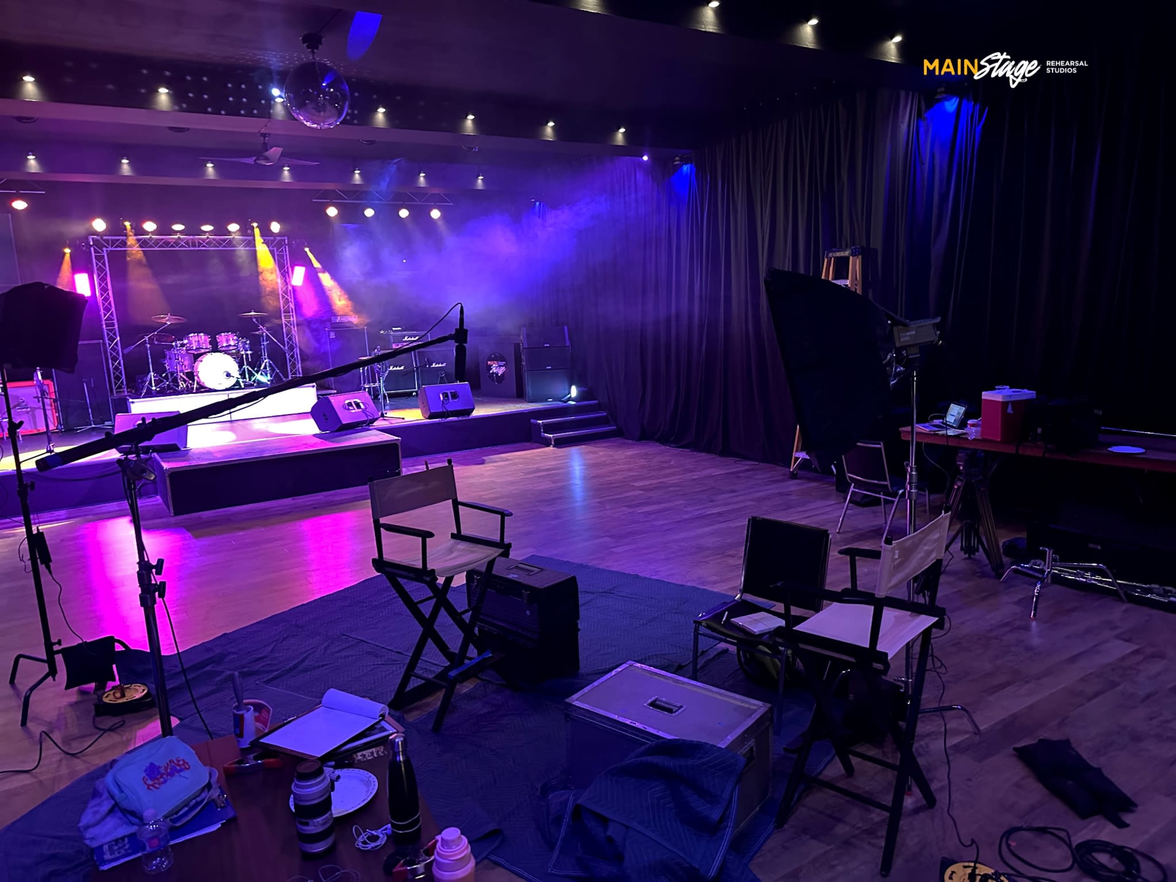 The image shows a concert venue stage set up with musical instruments, spotlights, and a seating area featuring director's chairs in a dimly lit space.