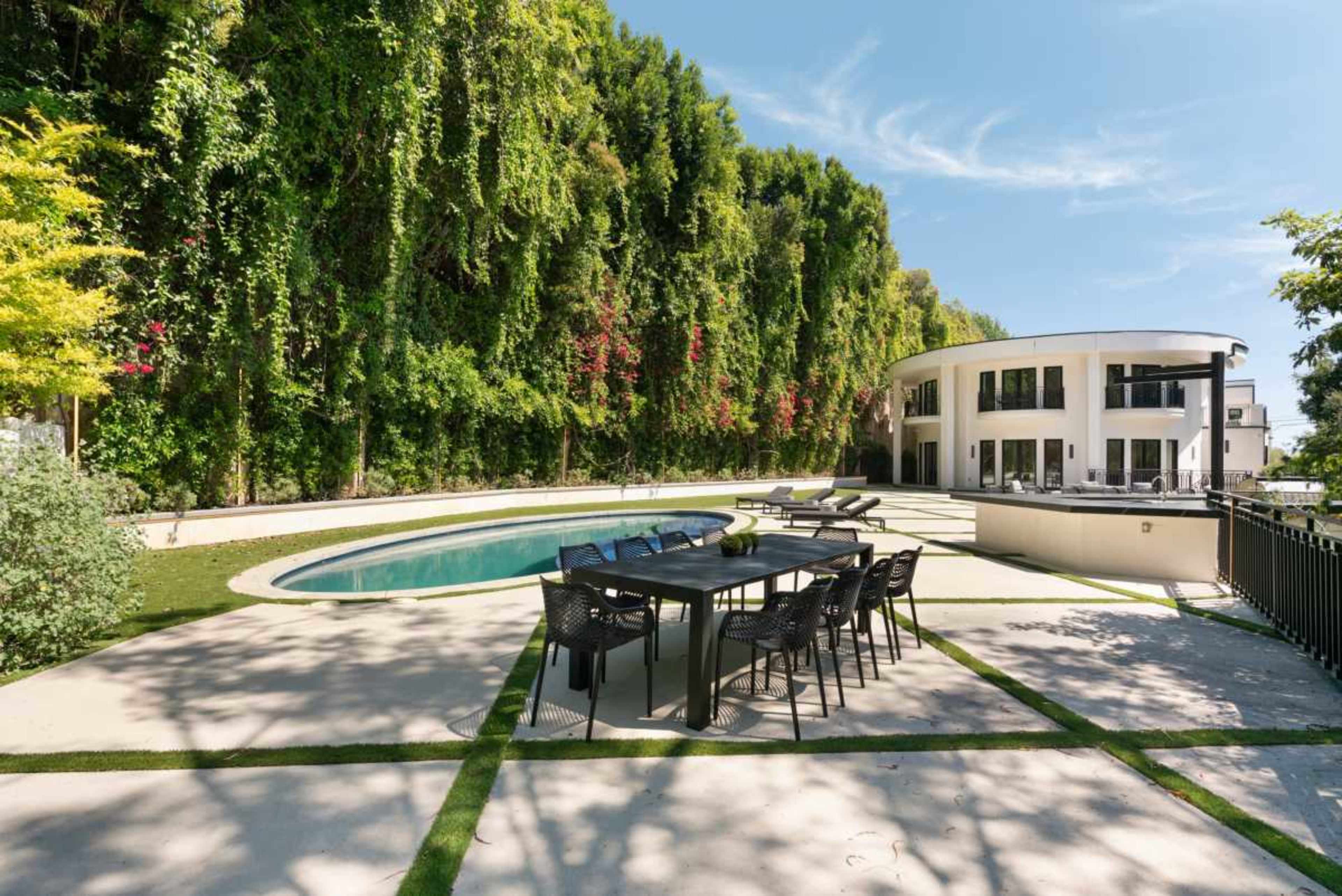 The image shows a modern house with a circular design situated next to a swimming pool and surrounded by lush greenery.