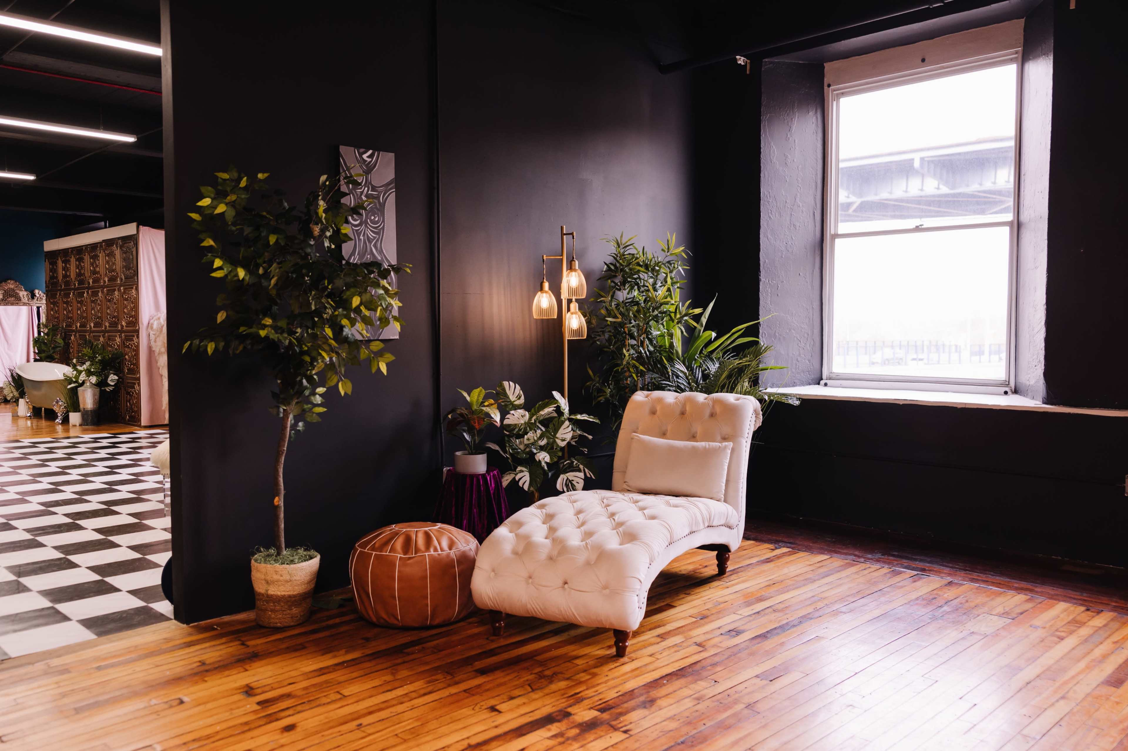 The image shows a chic corner of a room featuring a white chaise lounge, a floor lamp, potted plants, and hardwood flooring.