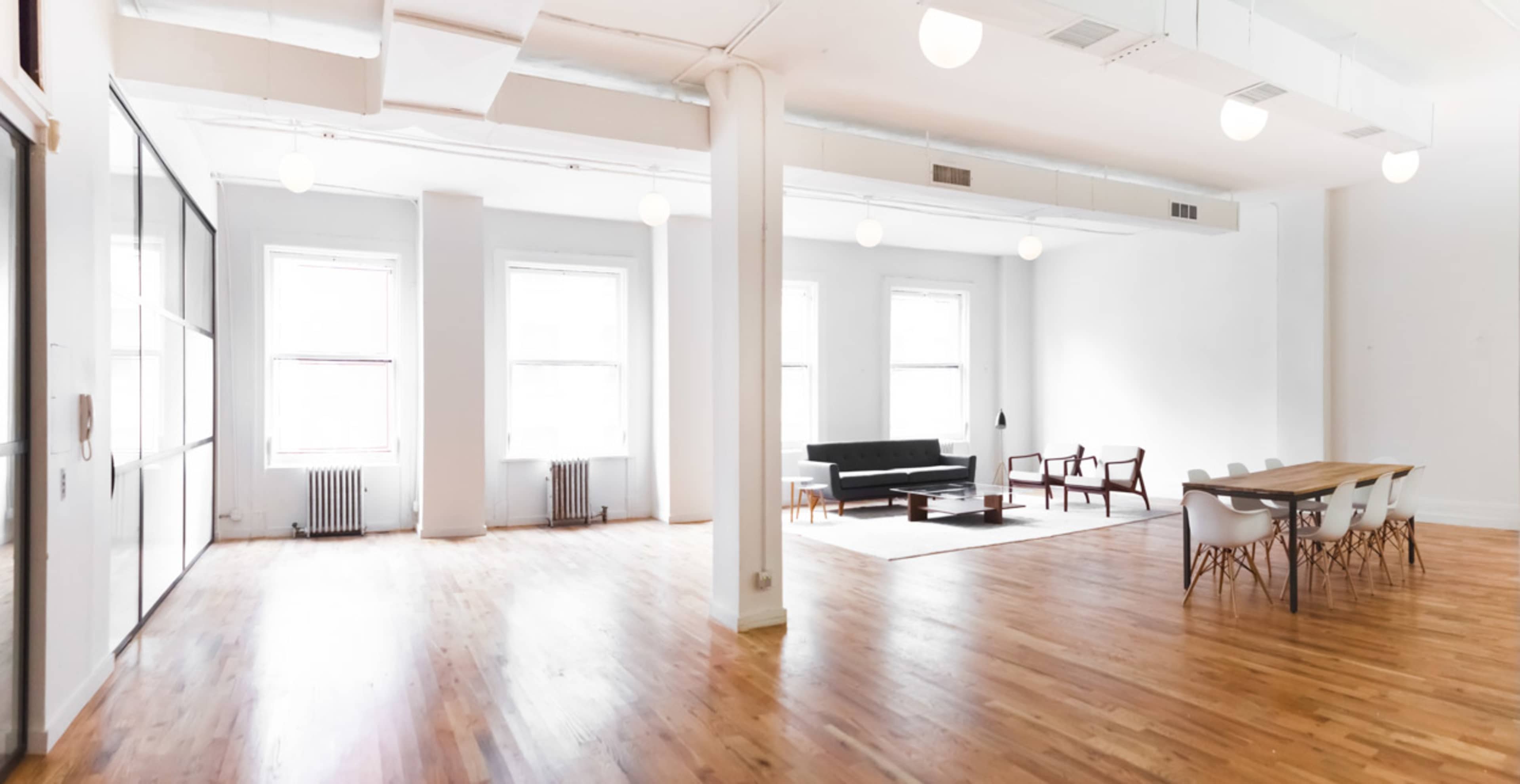 The image shows a spacious, open-plan room with wooden flooring, large windows, a seating area with modern furniture, and a dining table set for several people.