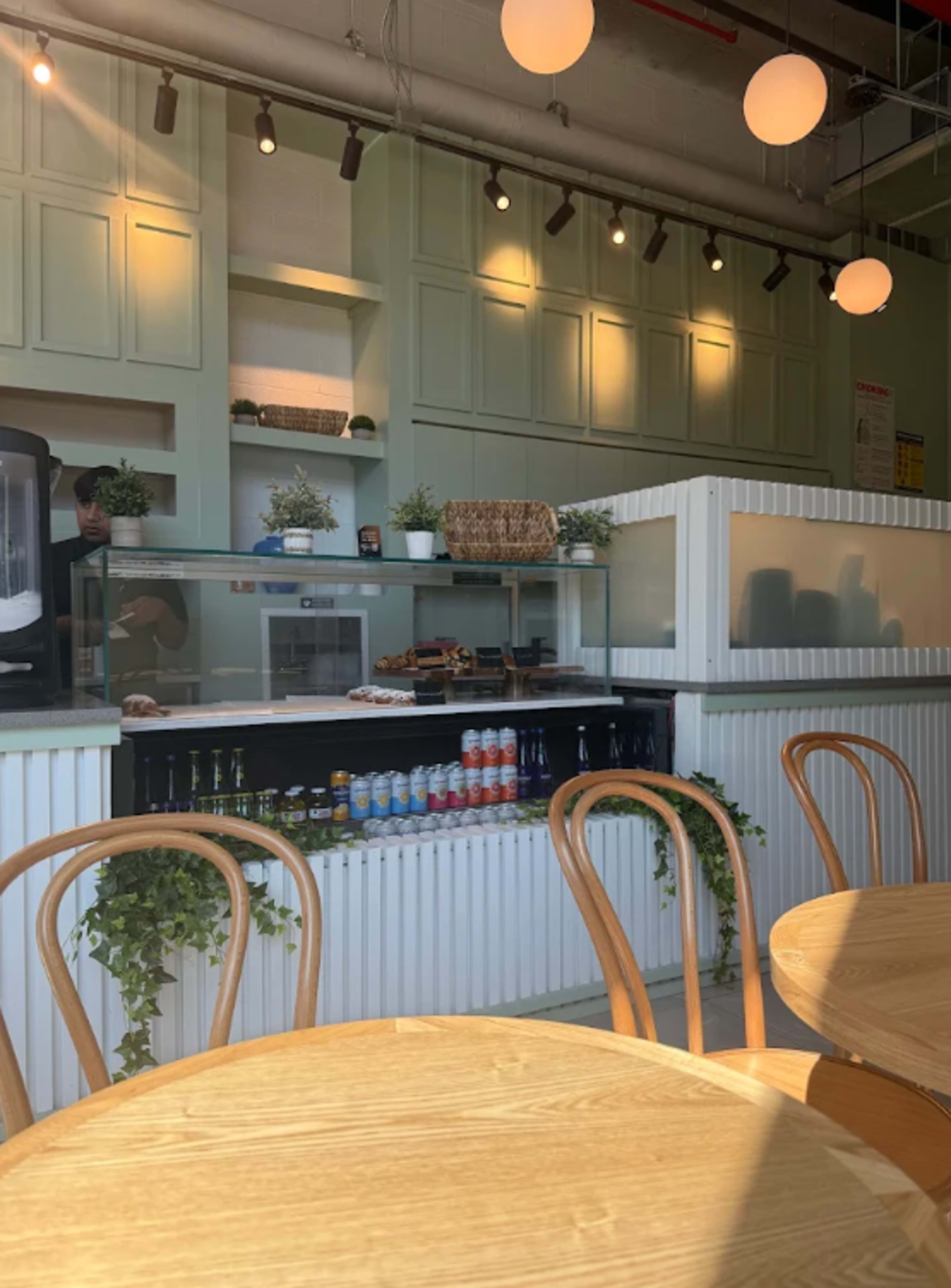 The image shows a modern café interior with a counter displaying various drinks and small plants, alongside wooden tables and minimalistic decor.