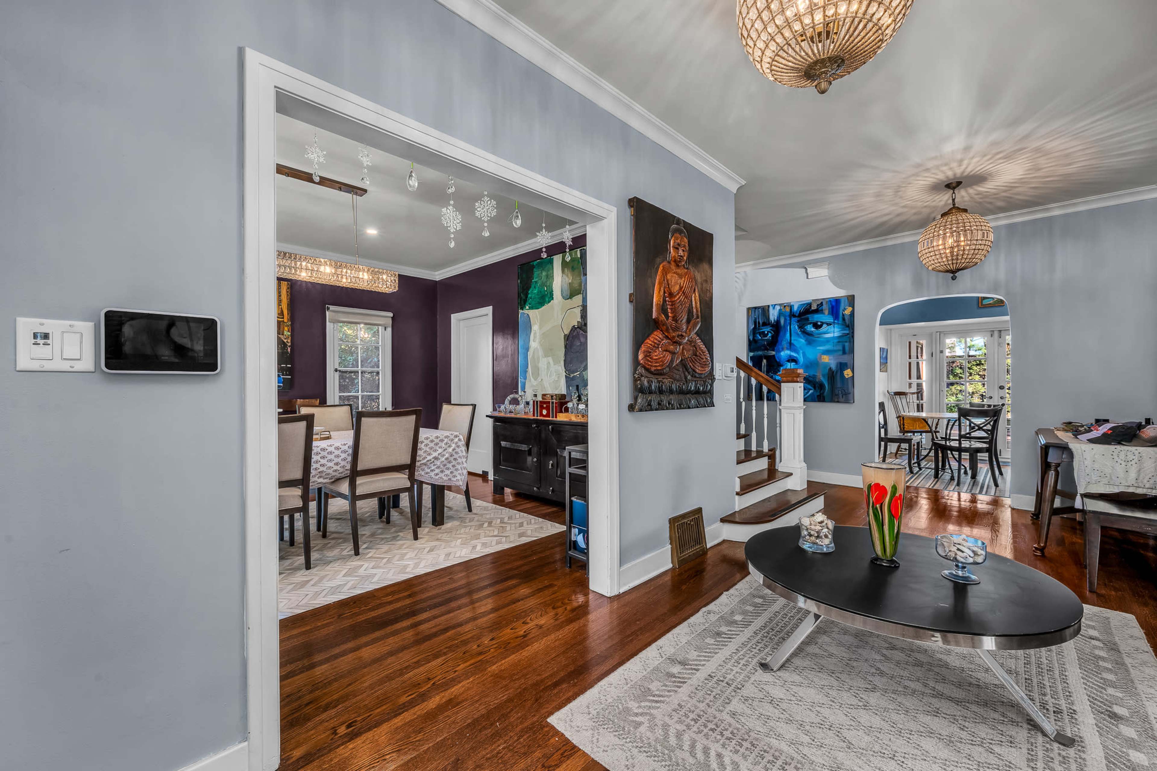 The image shows a spacious living area featuring a modern coffee table, decorative artwork on the walls, and a view into a dining room with a large table set for meals.