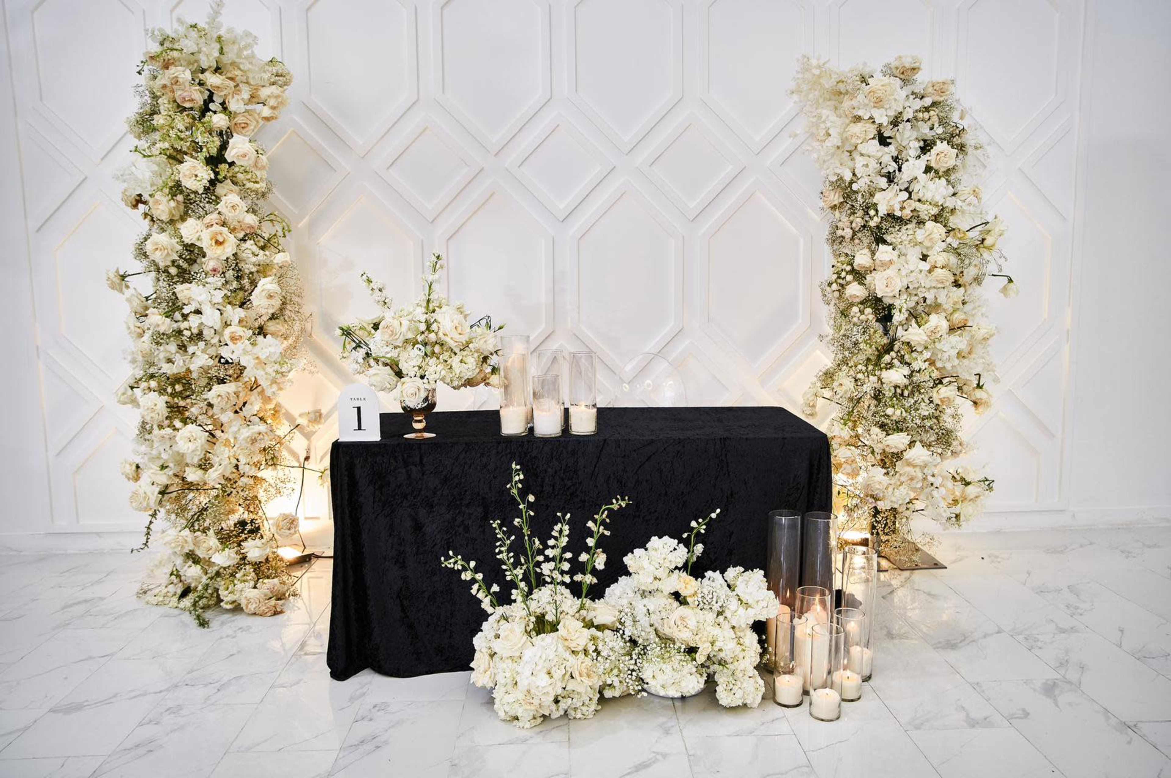 A black table is adorned with candles and floral arrangements, flanked by two tall floral displays against a textured white background.