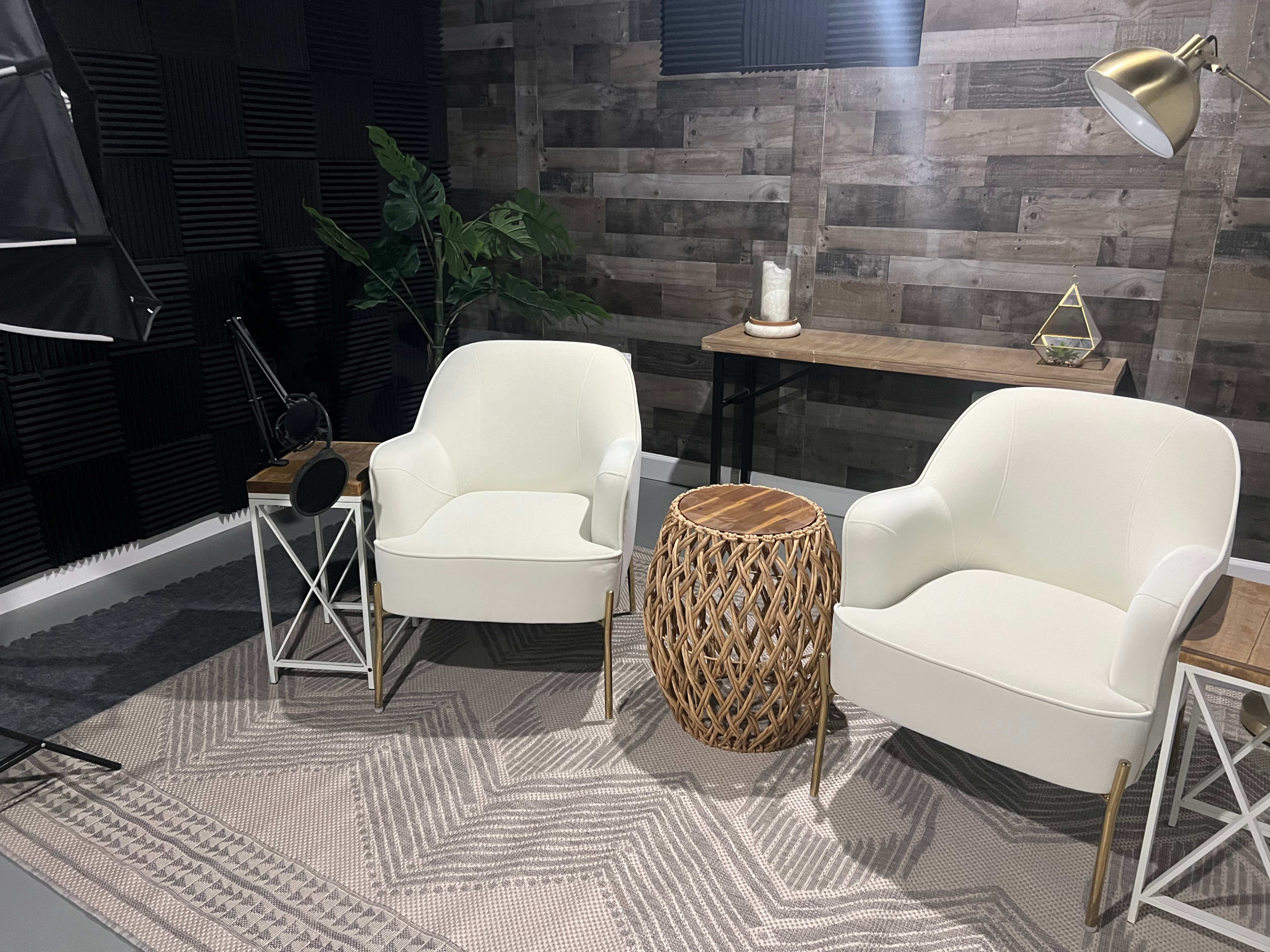 A cozy studio setup featuring two white chairs, a woven side table, and greenery against a wooden wall backdrop.