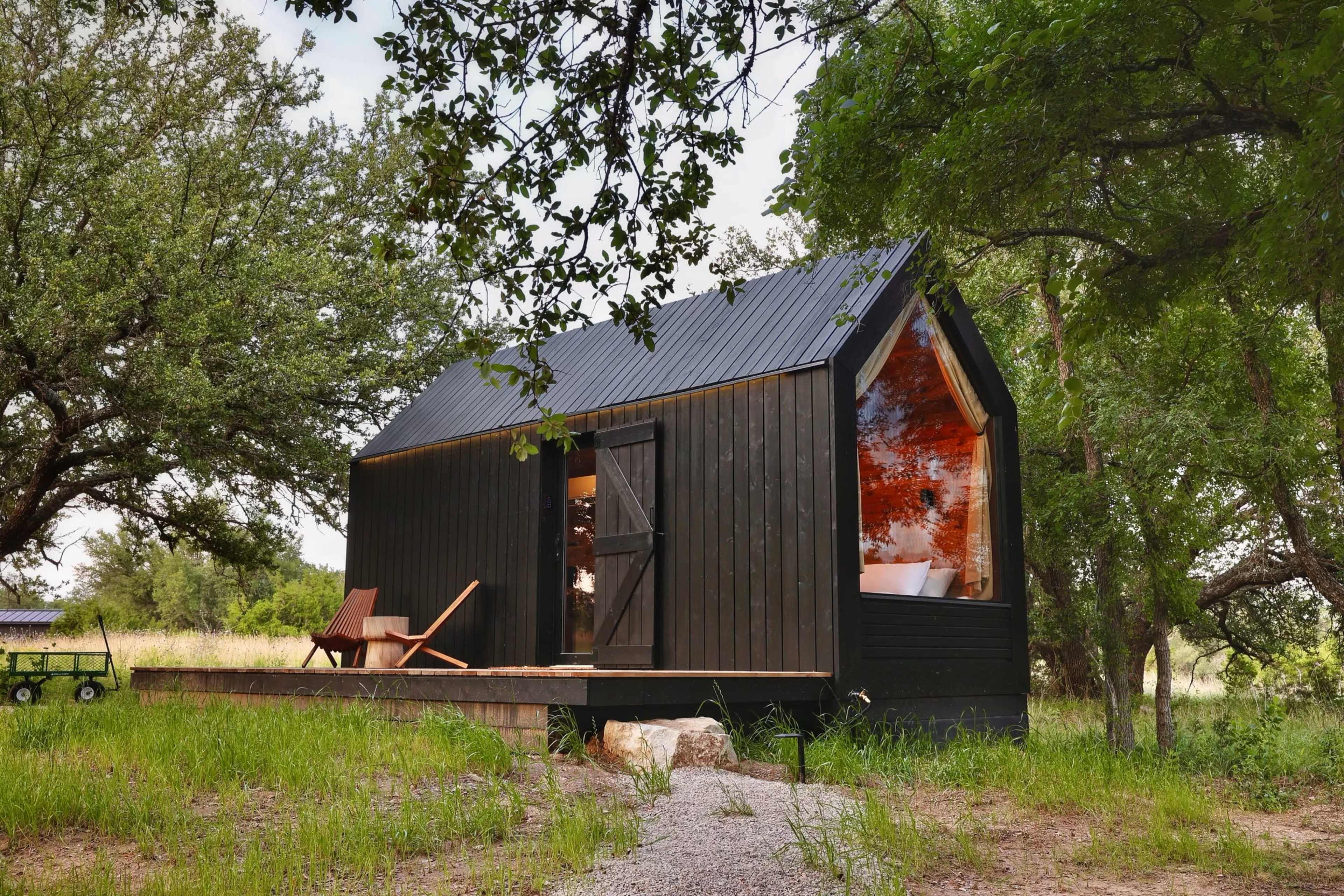 A small black wooden cabin with a metal roof sits surrounded by grass and trees, featuring large windows that reflect the landscape.