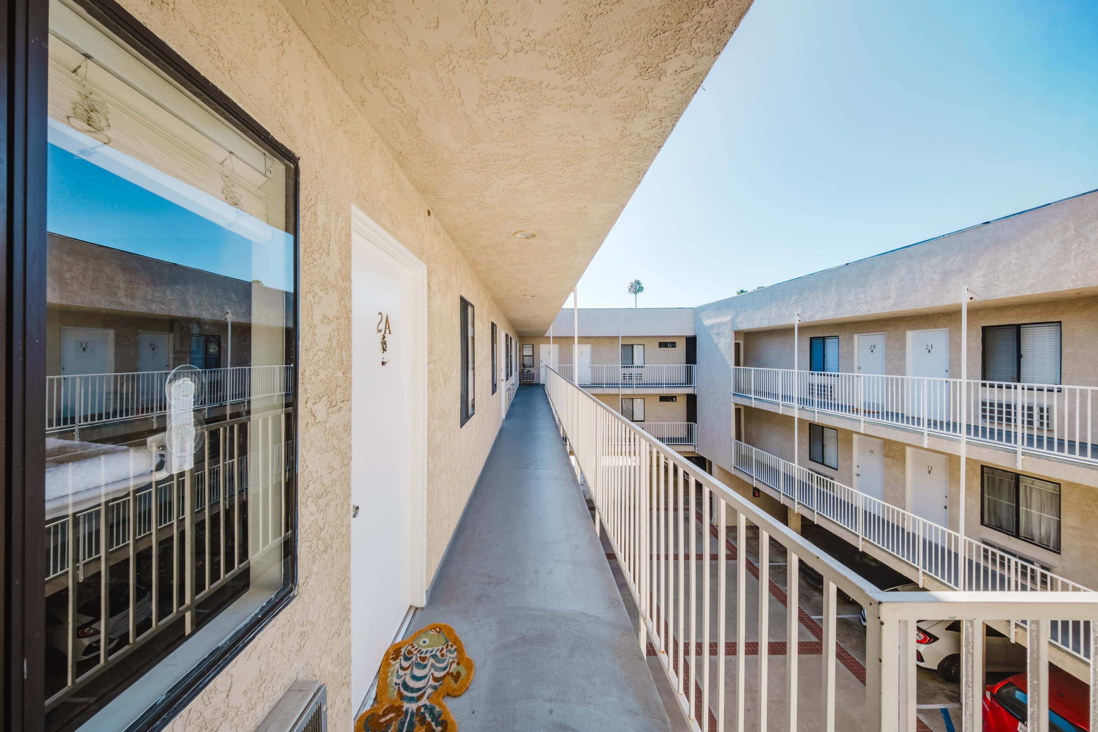 The image shows a long, clear walkway connecting rooms in a two-story motel with a view of parked cars and a blue sky.