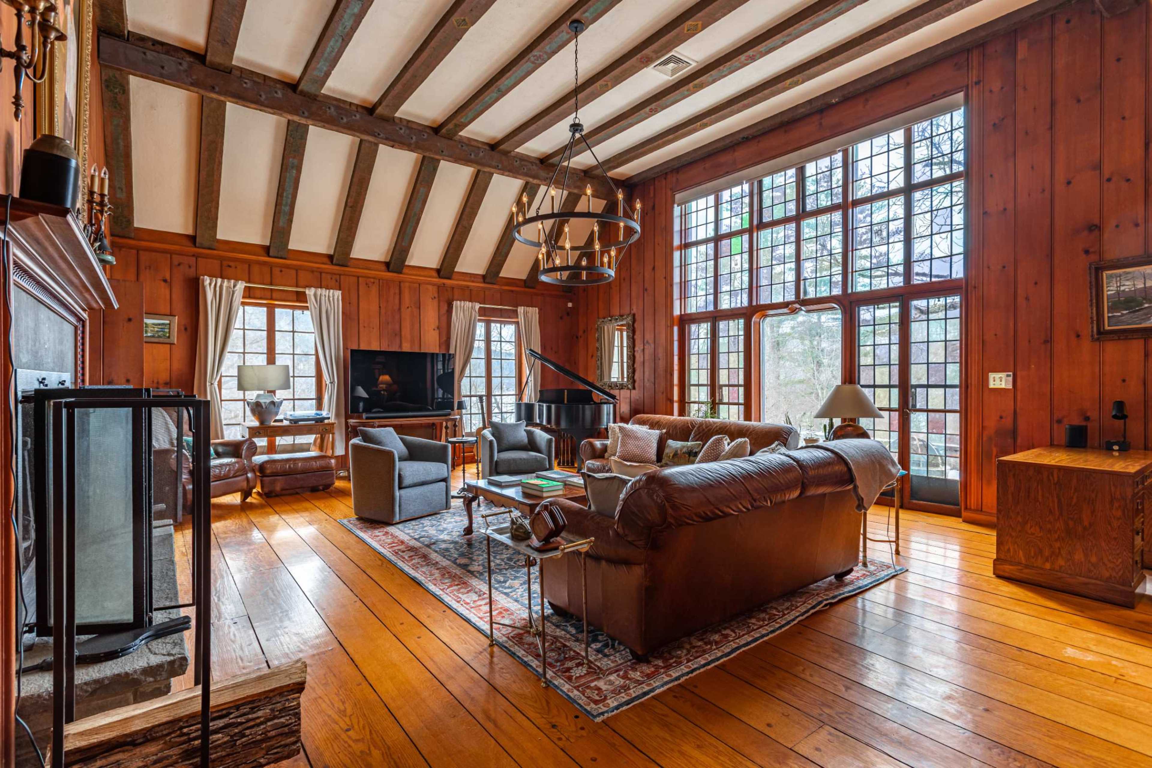 The spacious living room features wooden walls, a high beamed ceiling, large windows, and a central seating arrangement around a fireplace.