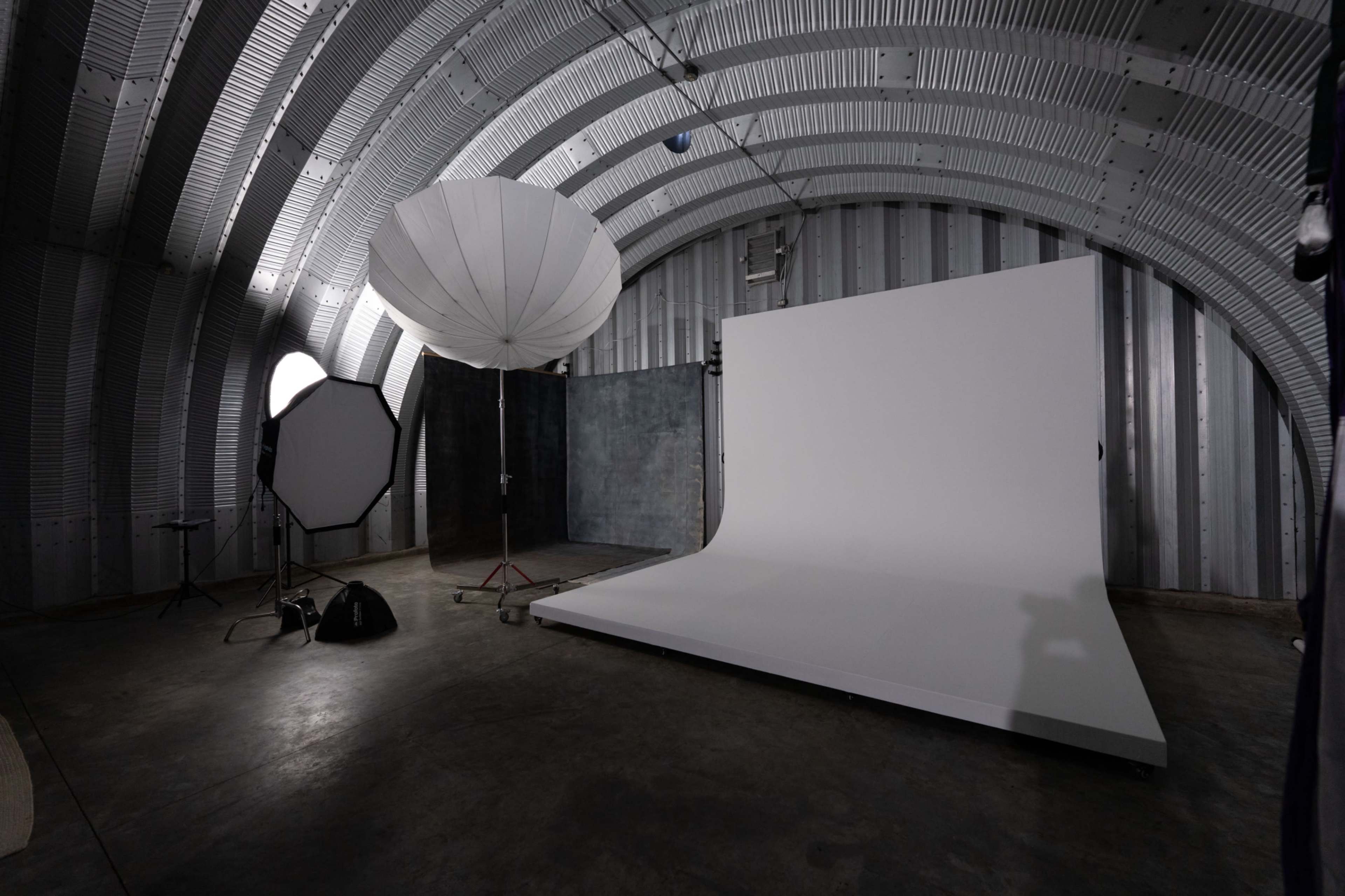 The image shows a photography studio featuring a large seamless backdrop, a reflector umbrella, and various lighting equipment arranged in an industrial space.