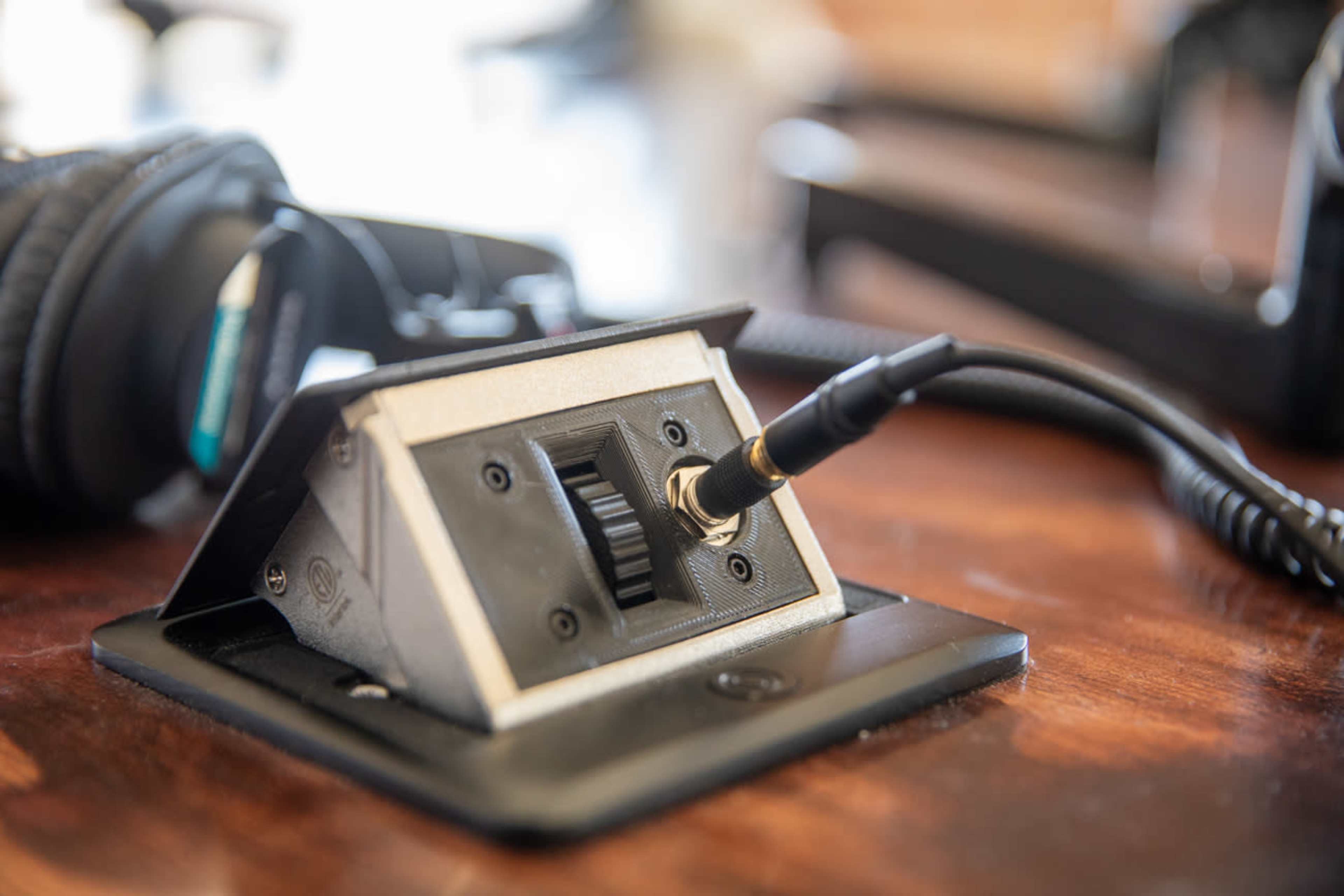 A close-up view of a headphone jack connected to a control panel on a wooden surface, with headphones partially visible in the background.