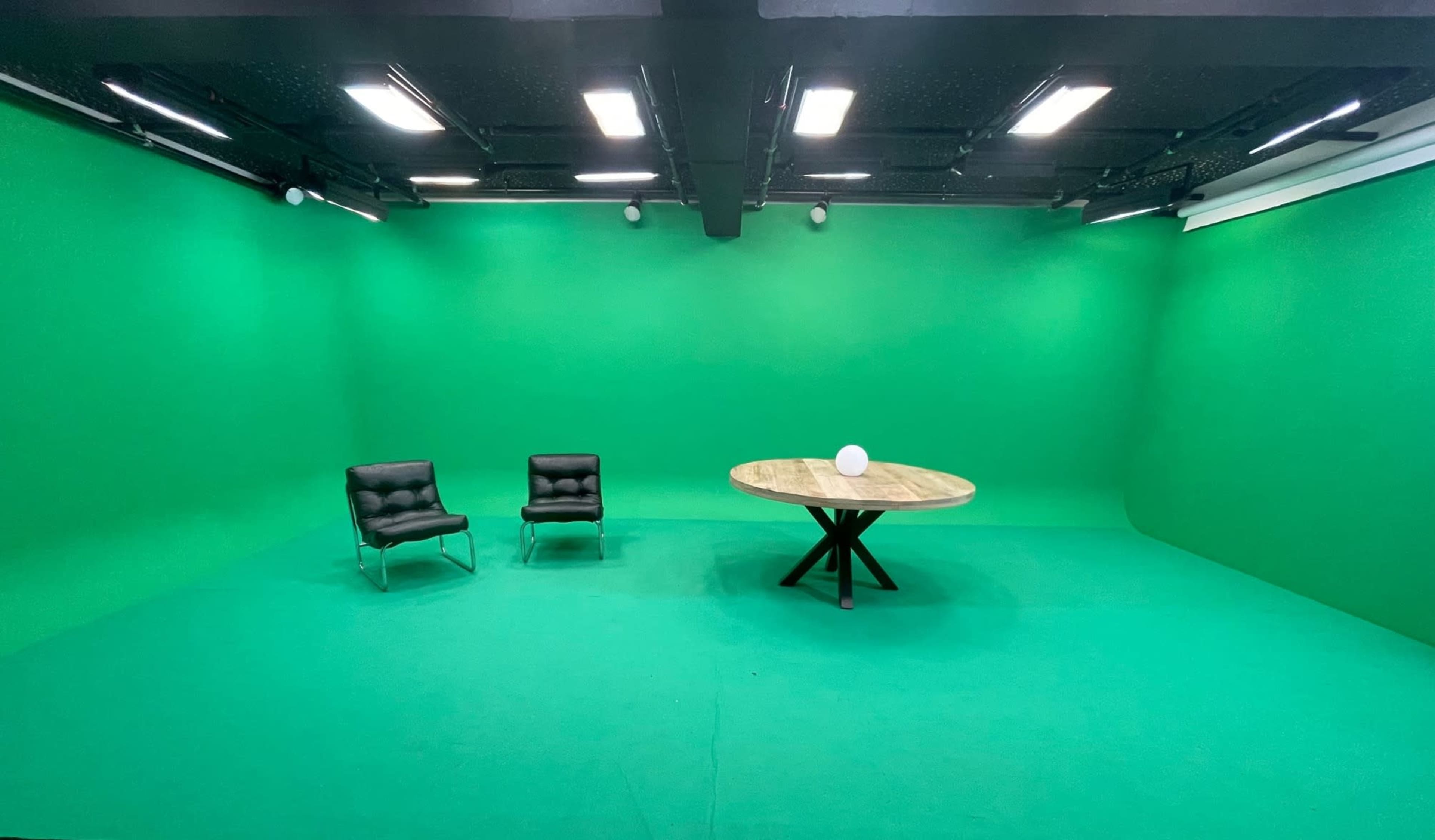 A green-screen studio with two black chairs and a round wooden table in the center.